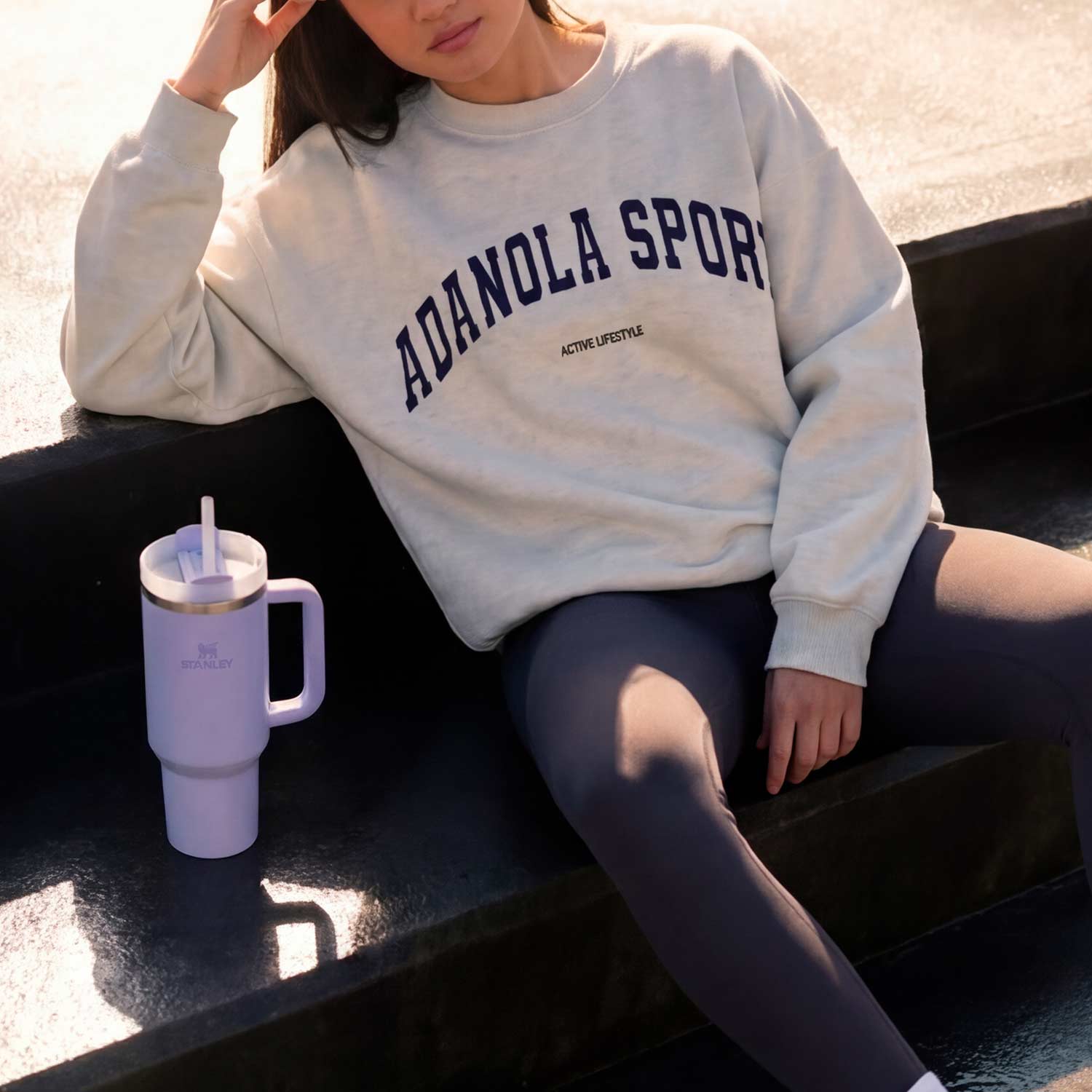 Person seated on steps with a 'ADANOLA SPORT' sweatshirt and purple mug with a straw beside them.