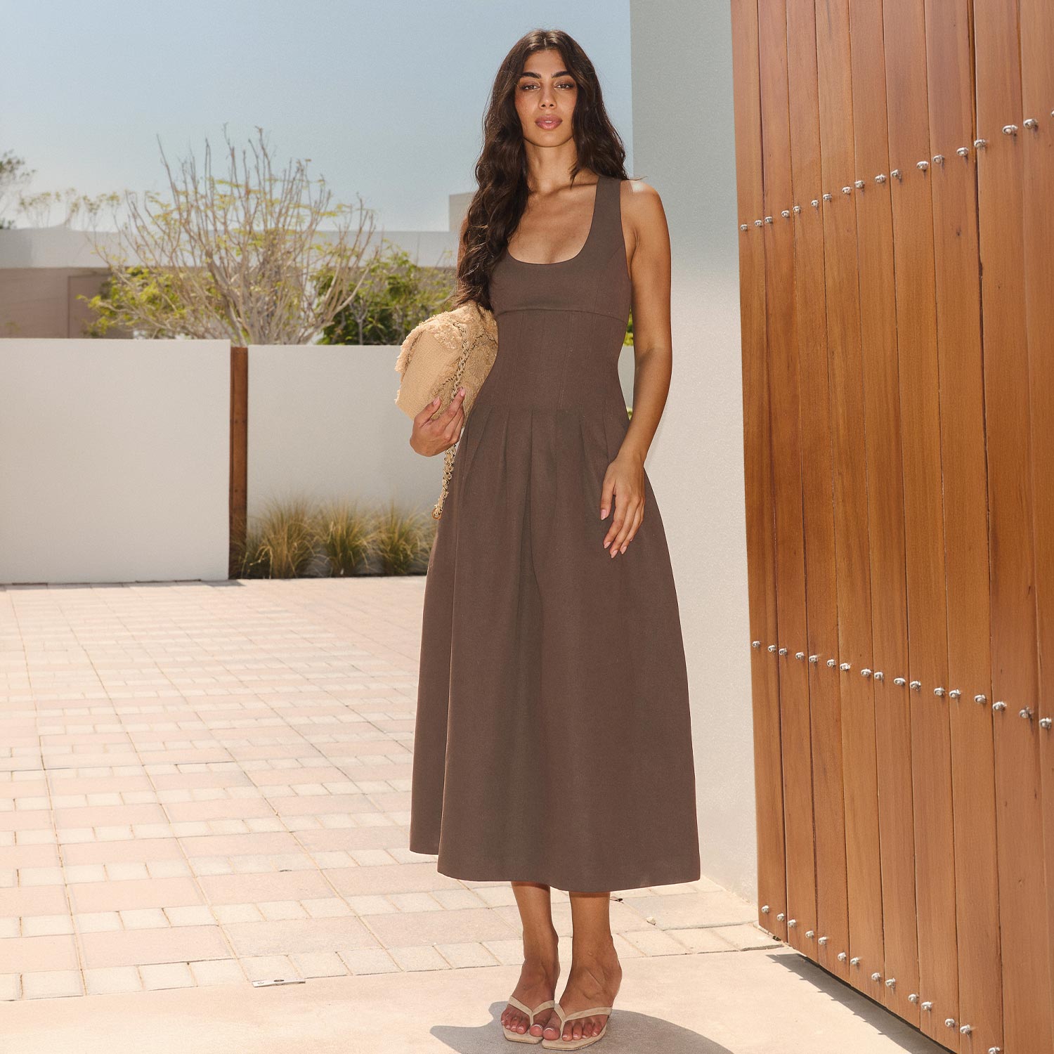 Woman wearing brown sleeveless midi dress with beige clutch bag, standing on terrace with wooden gate