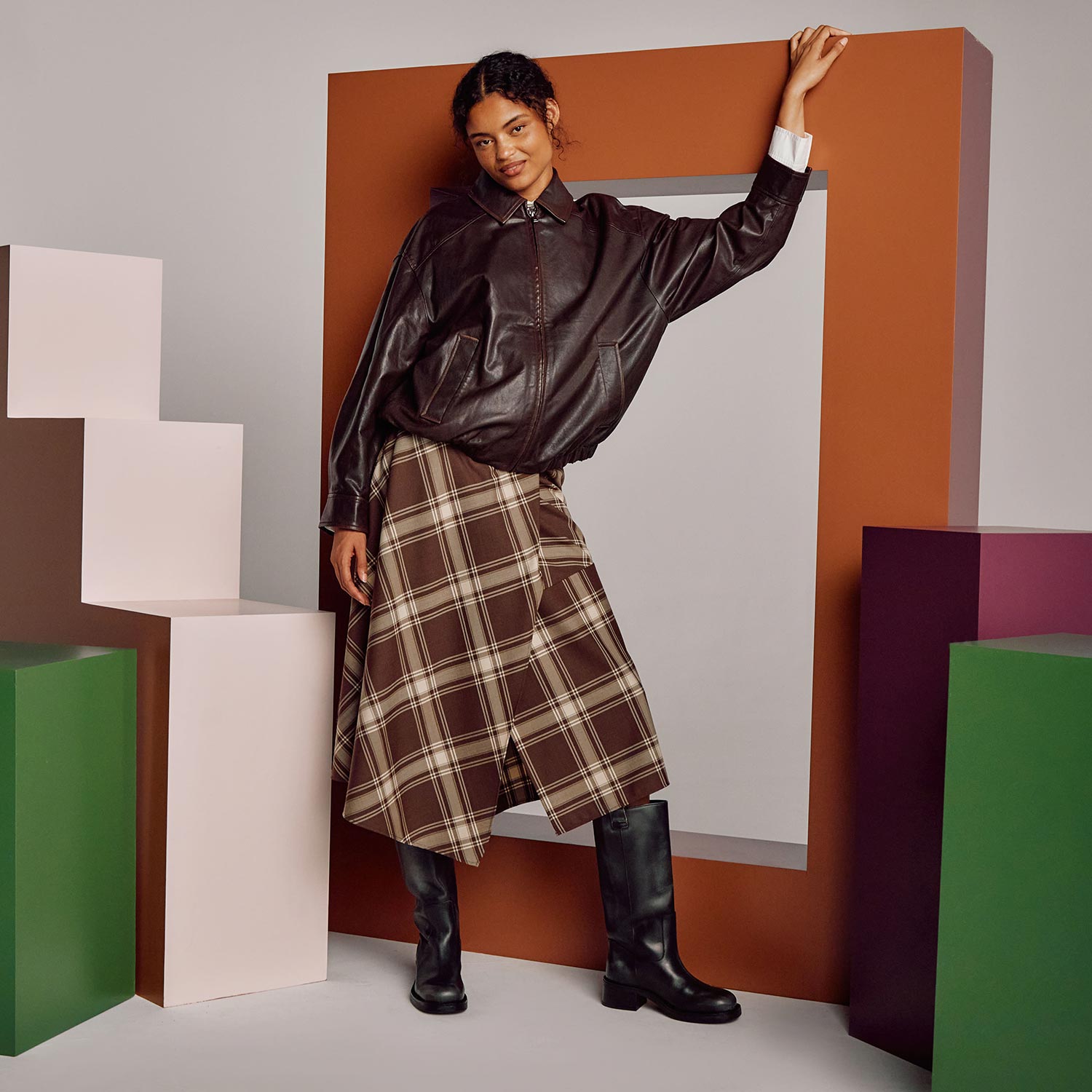 Woman wearing black leather jacket and brown tartan check midi skirt with black knee-high boots
