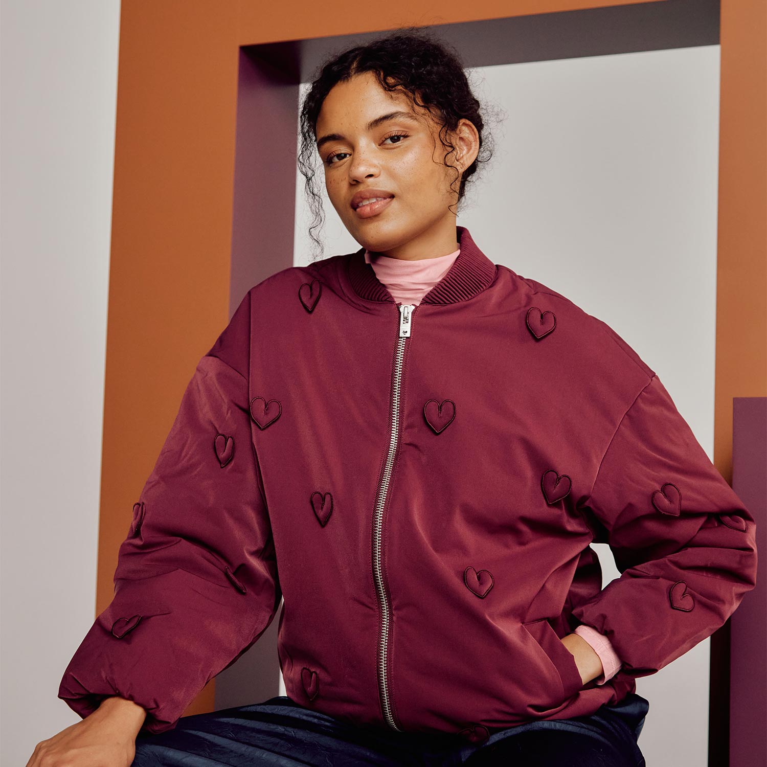 Woman wearing burgundy padded jacket with embroidered heart pattern and ribbed collar against orange and grey background