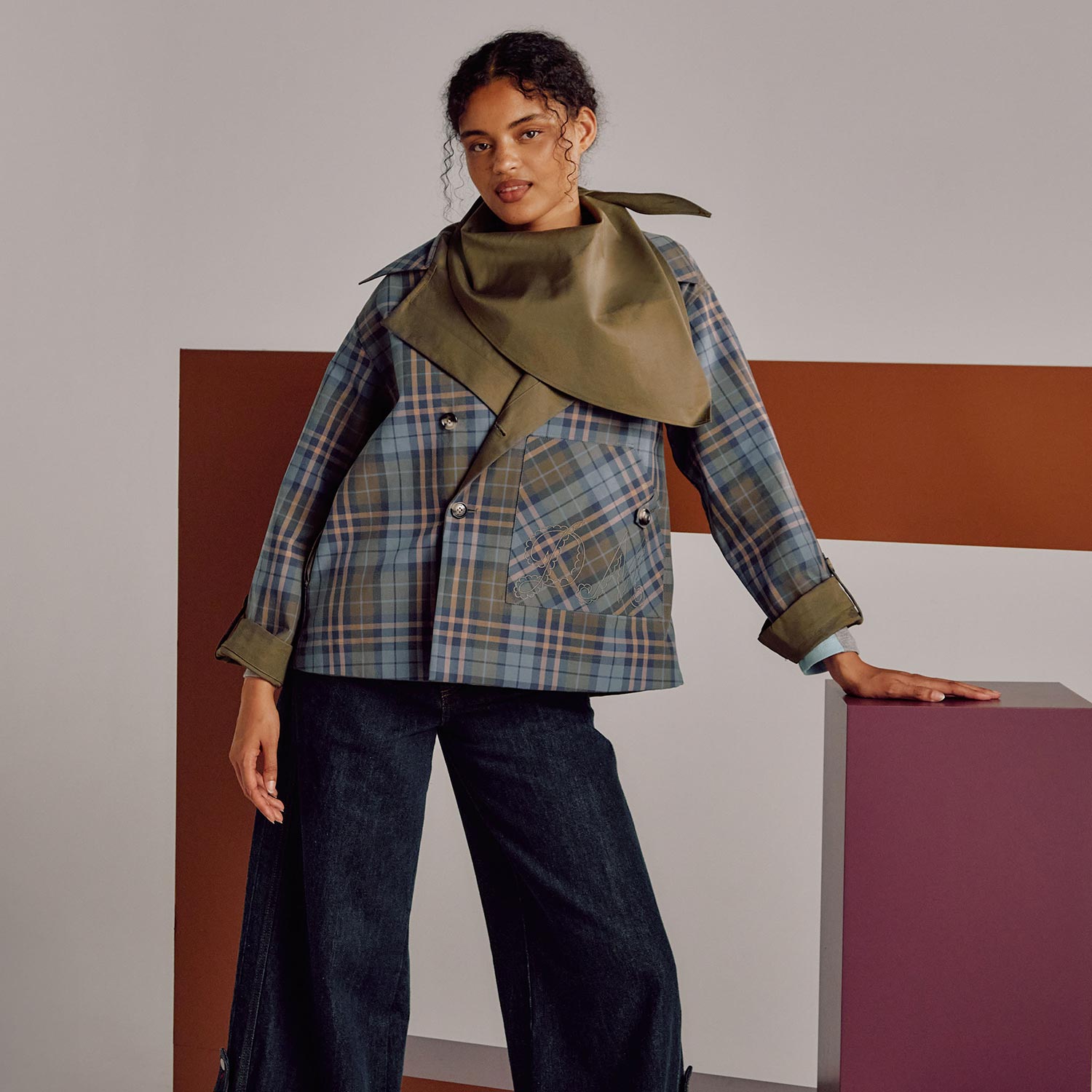 Woman wearing blue checked blazer with olive green collar detail and dark denim jeans against geometric background