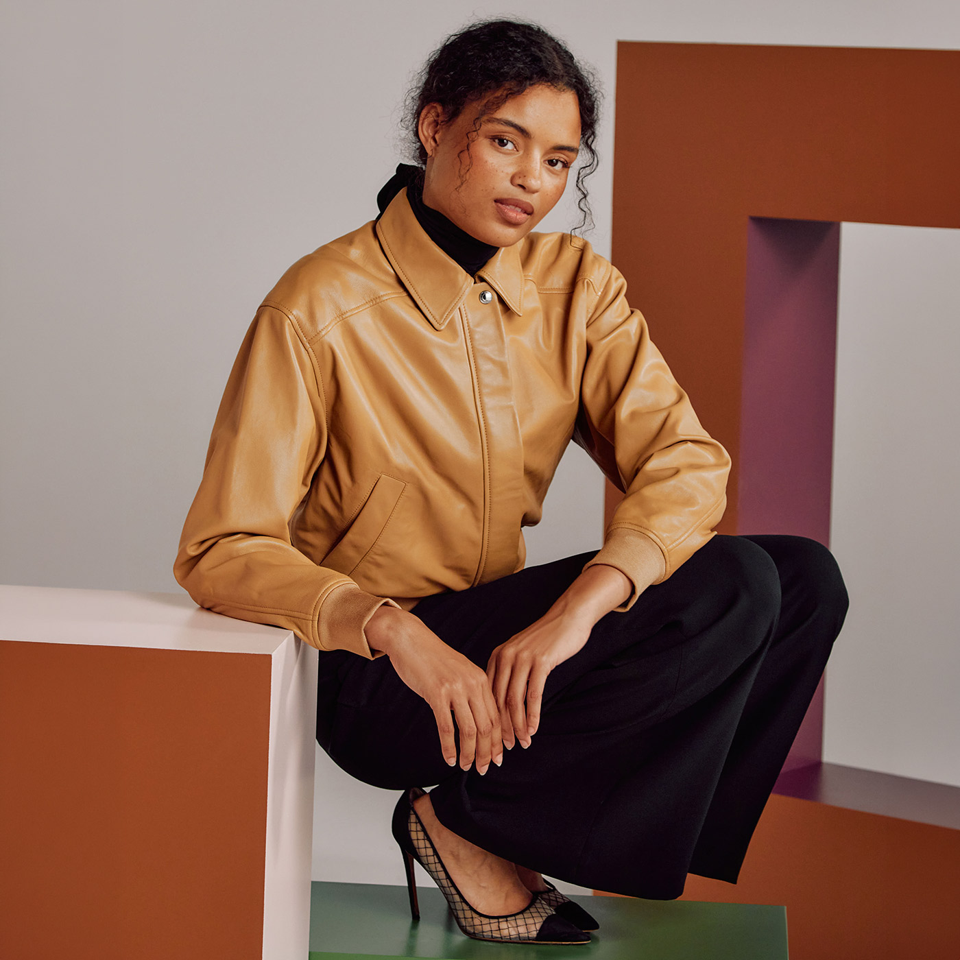 Woman wearing tan leather jacket and black trousers with mesh heels, seated against geometric background