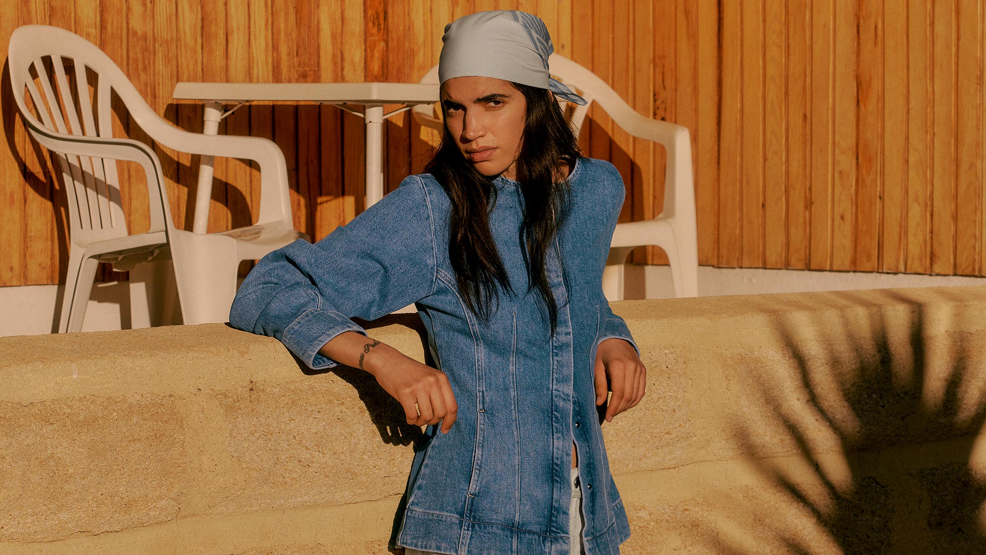 Woman in denim shirt dress and grey headscarf posing outdoors against wooden backdrop