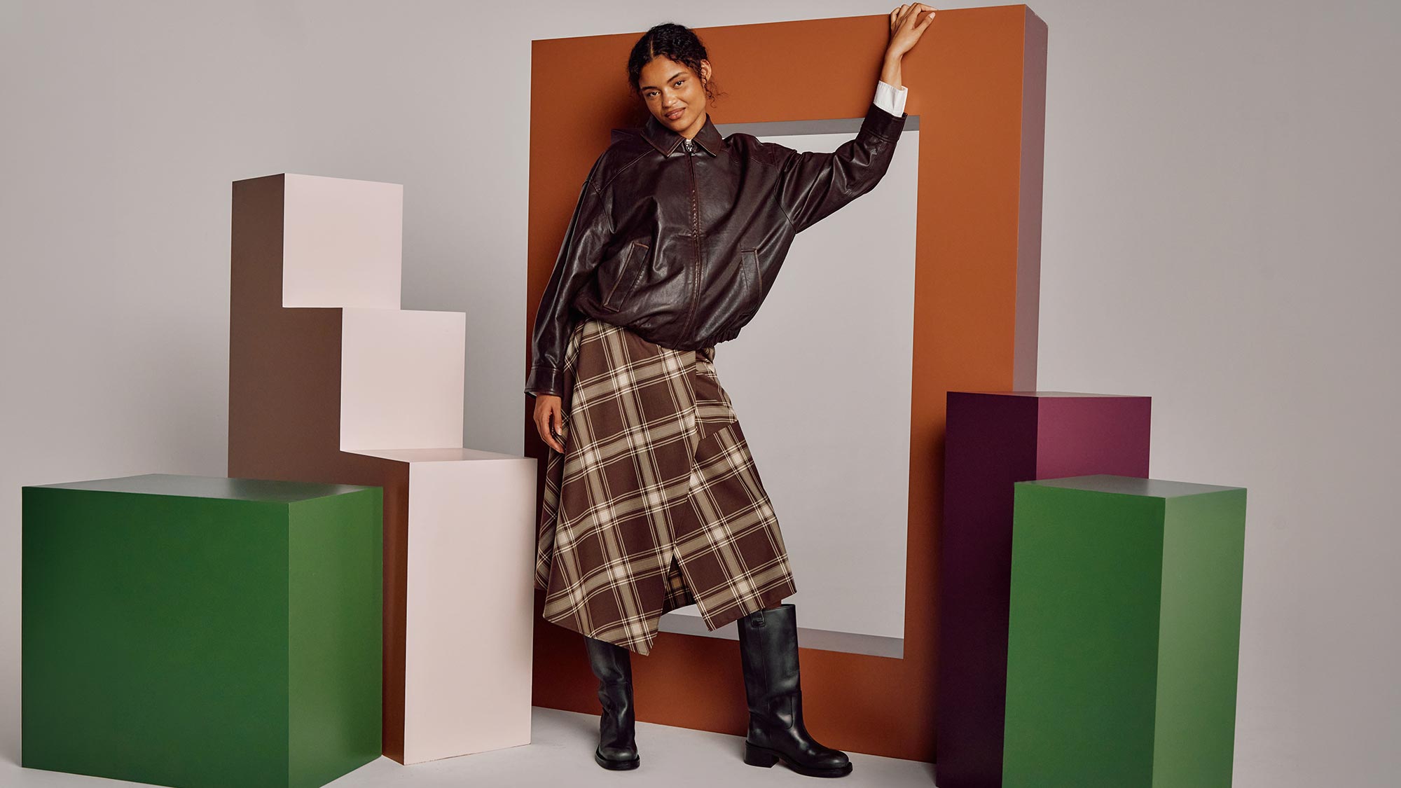 Woman in black leather jacket and brown tartan midi skirt with black boots, posing against colourful geometric backdrop