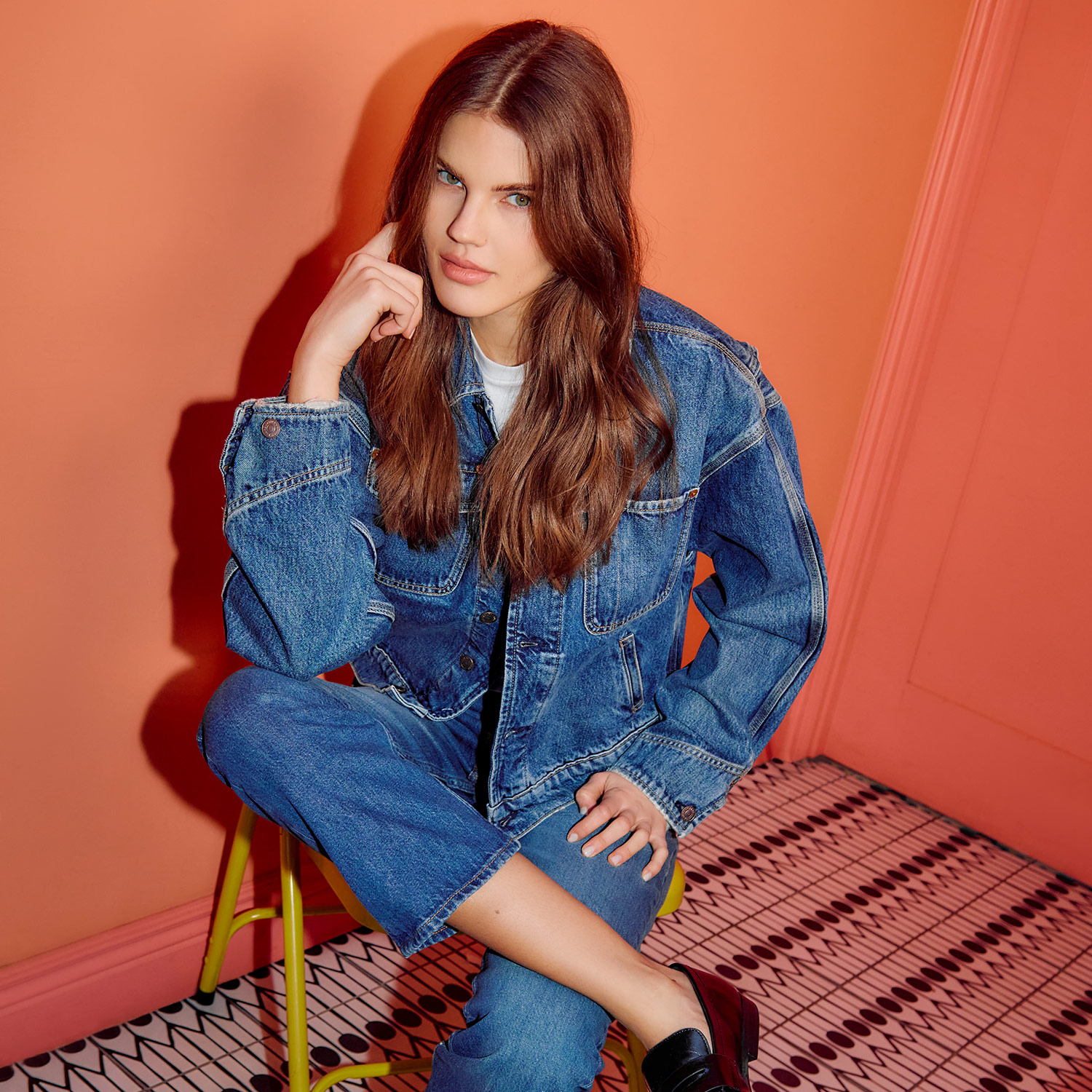 A person with long brown hair wearing a denim jacket poses against an orange background.