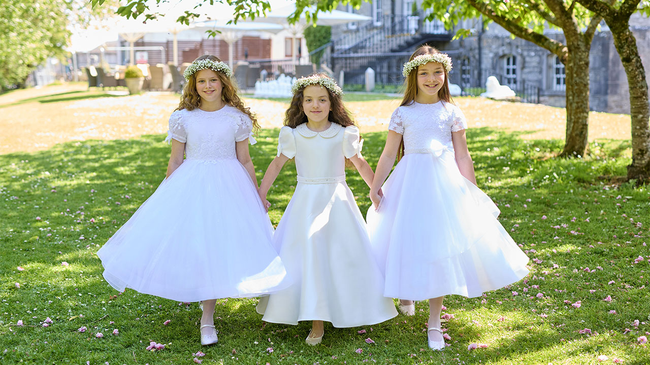 Three young girls in white dresses and floral headbands walk hand in hand on a sunlit grassy area.
