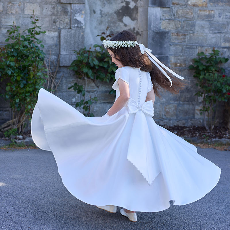 Young girl in a white dress with a floral headband twirls outside near a stone wall and greenery.