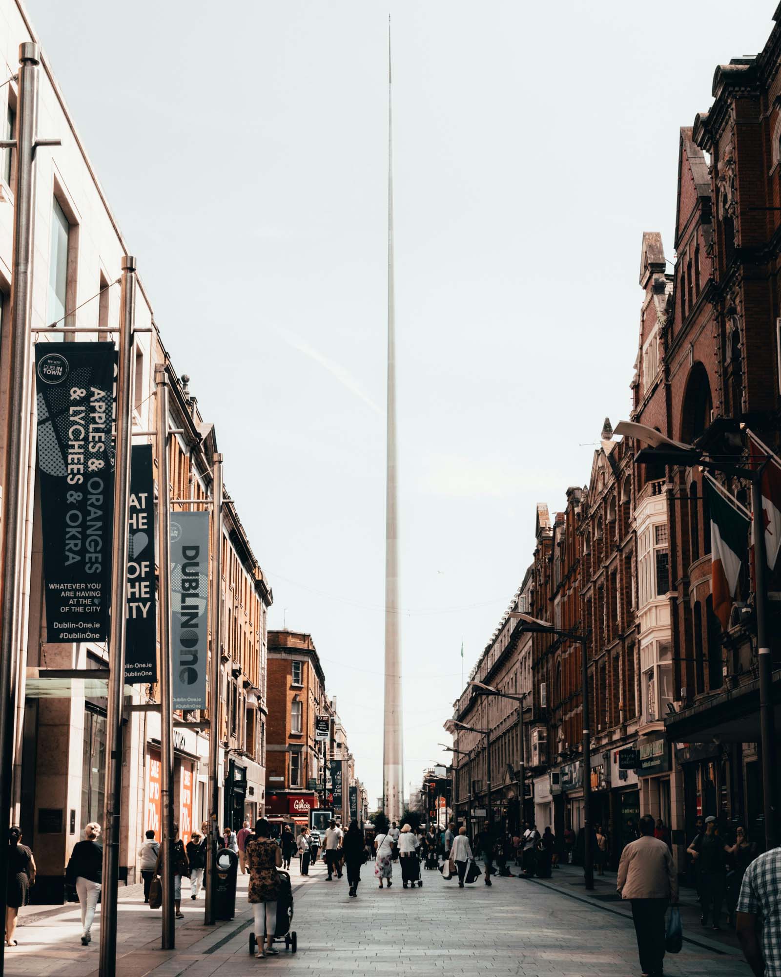 A bustling street with people walking, flanked by buildings on both sides, and a tall spire in the distance under a clear sky.