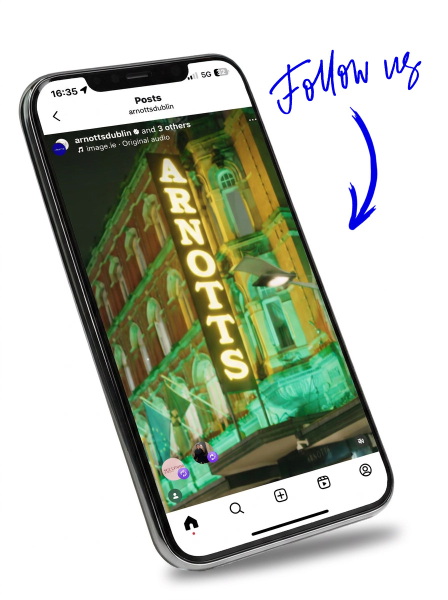 A smartphone displaying an Instagram post featuring the illuminated Arnotts sign on a historic building at night. The building is lit with green and yellow lights. Above the phone, blue text reads 'Follow us' with an arrow pointing downwards.