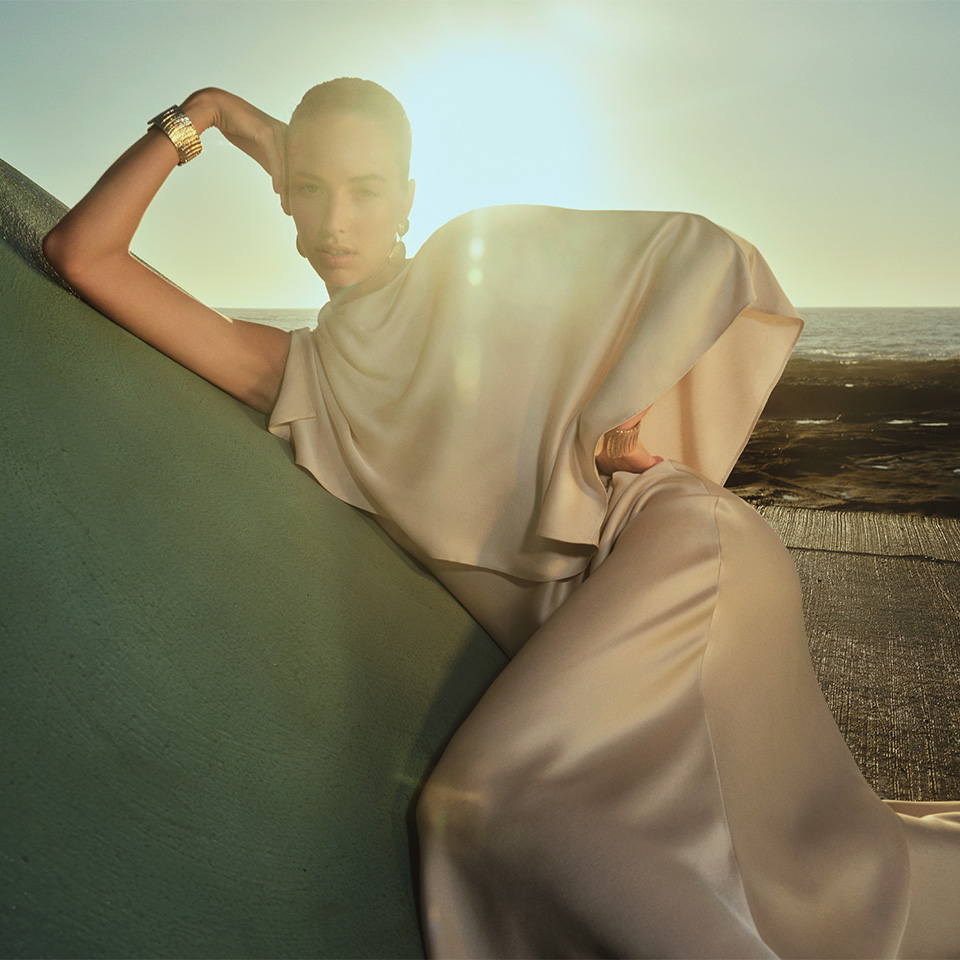 A woman in an elegant cream dress poses outdoors, leaning on a green surface with sunlight in the background.
