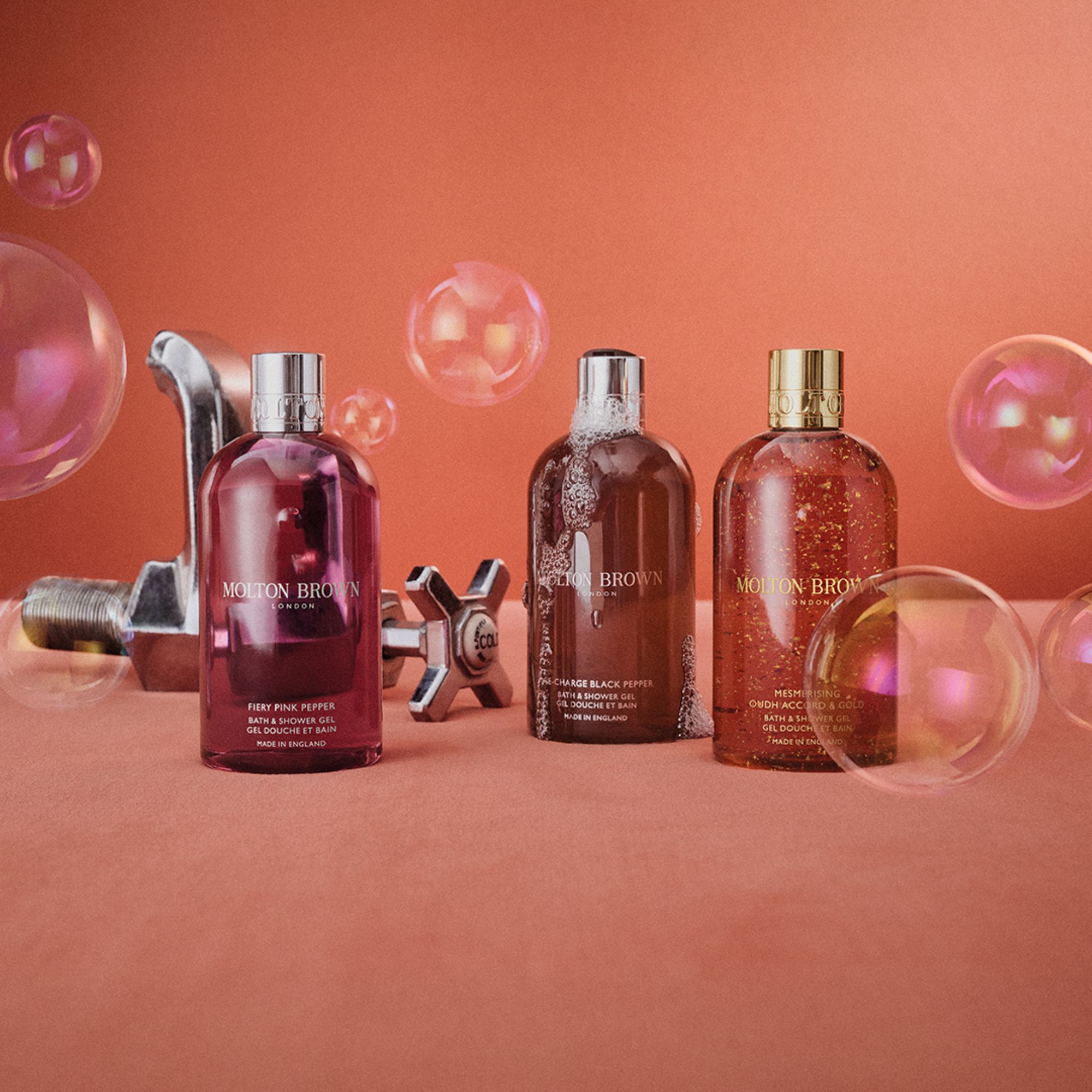 Molton Brown bath and body products including body washes and shower gels in jewel-toned bottles with decorative spheres on coral background