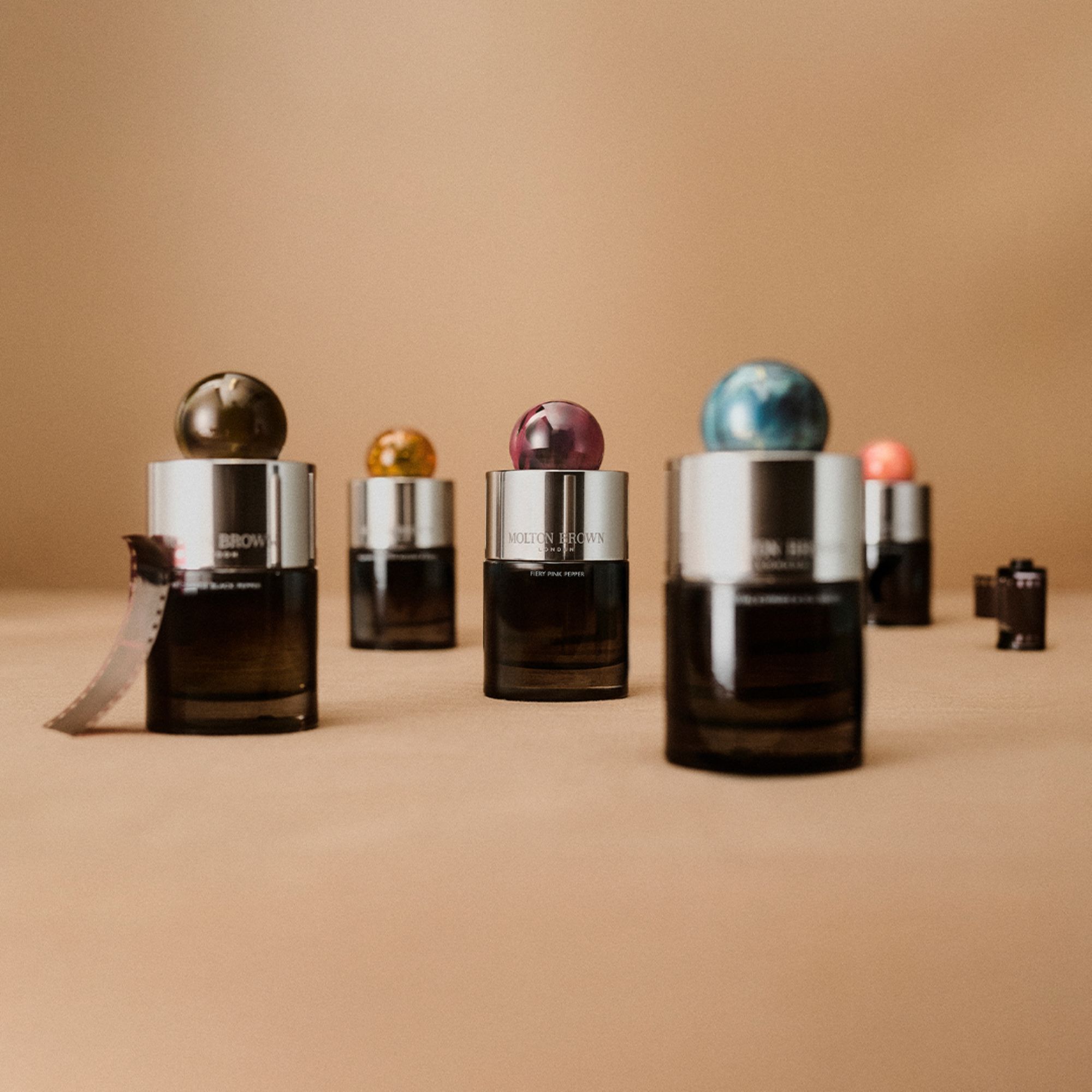 Molton Brown fragrance bottles with metallic sphere tops in various colours displayed on neutral beige background