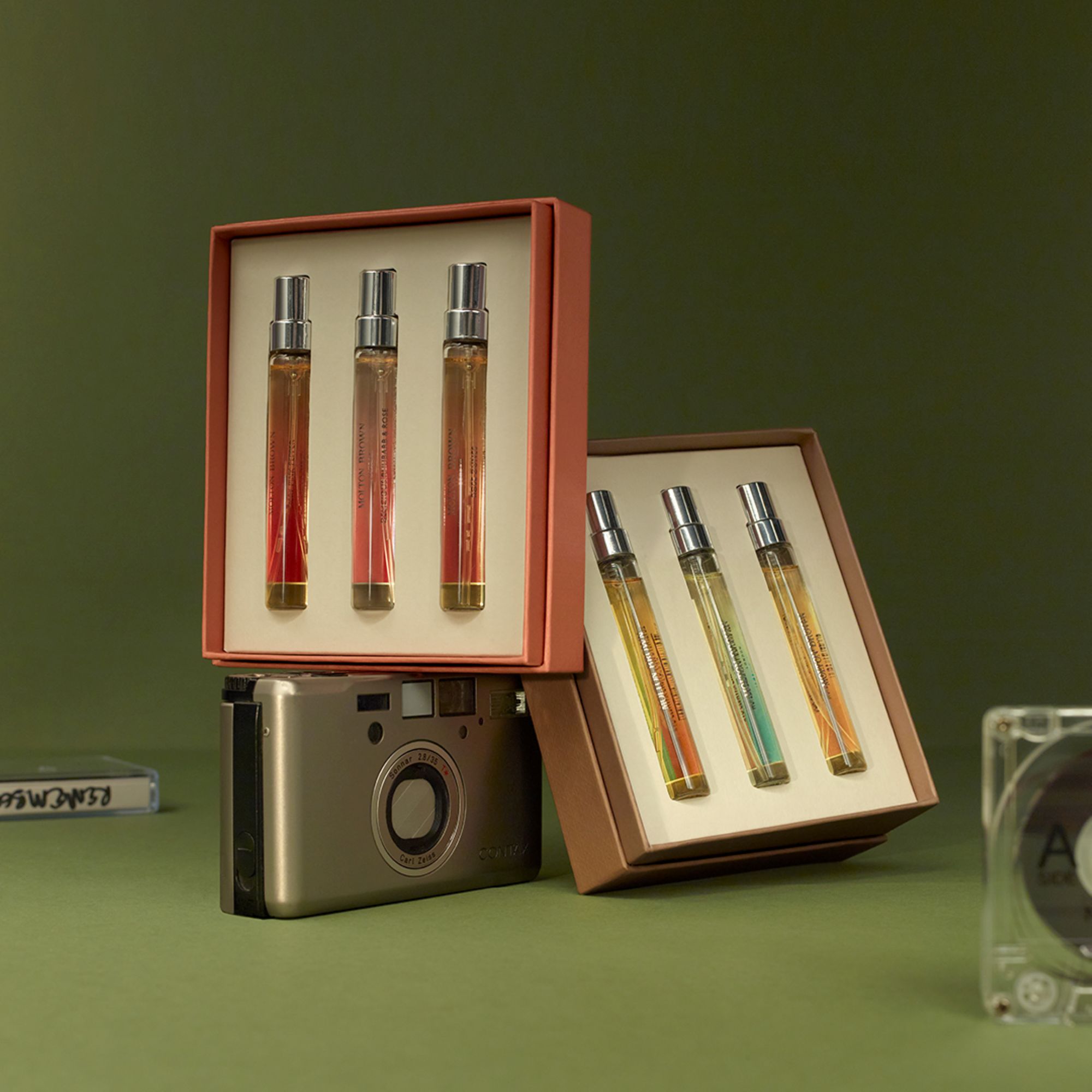 Molton Brown fragrance gift sets in coral and tan boxes containing spray bottles, displayed with vintage camera on green background