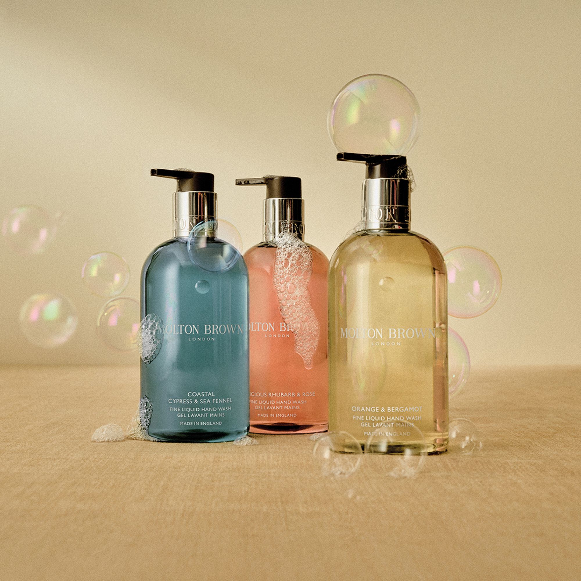 Three Molton Brown fine liquid hand wash bottles in blue, coral, and amber with pump dispensers and decorative bubbles