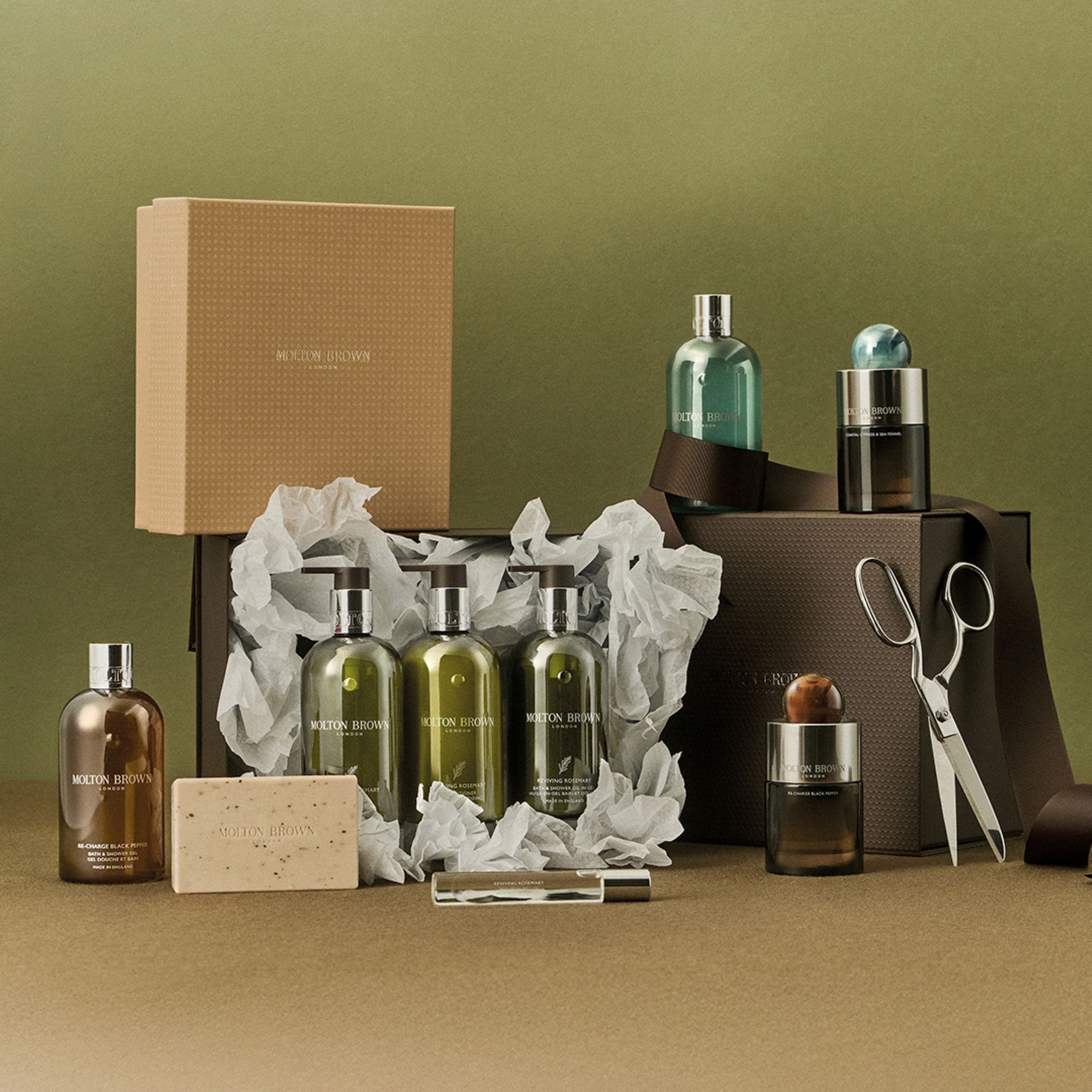 Molton Brown home fragrance gift set with multiple bottles, candles, soap, scissors, and kraft packaging displayed against olive green background
