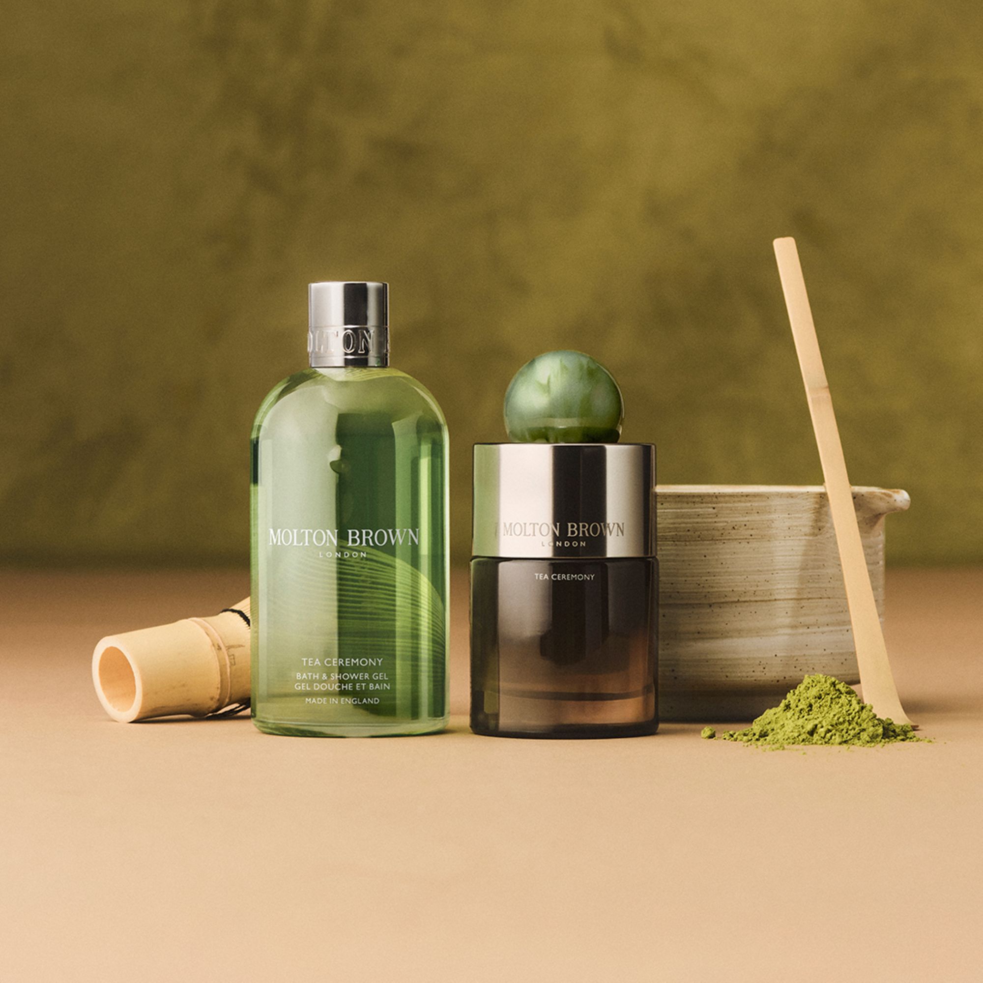 Molton Brown Tea Ceremony collection featuring green bath and shower gel bottle, scented candle with green matcha powder, wooden whisk, and natural beige background