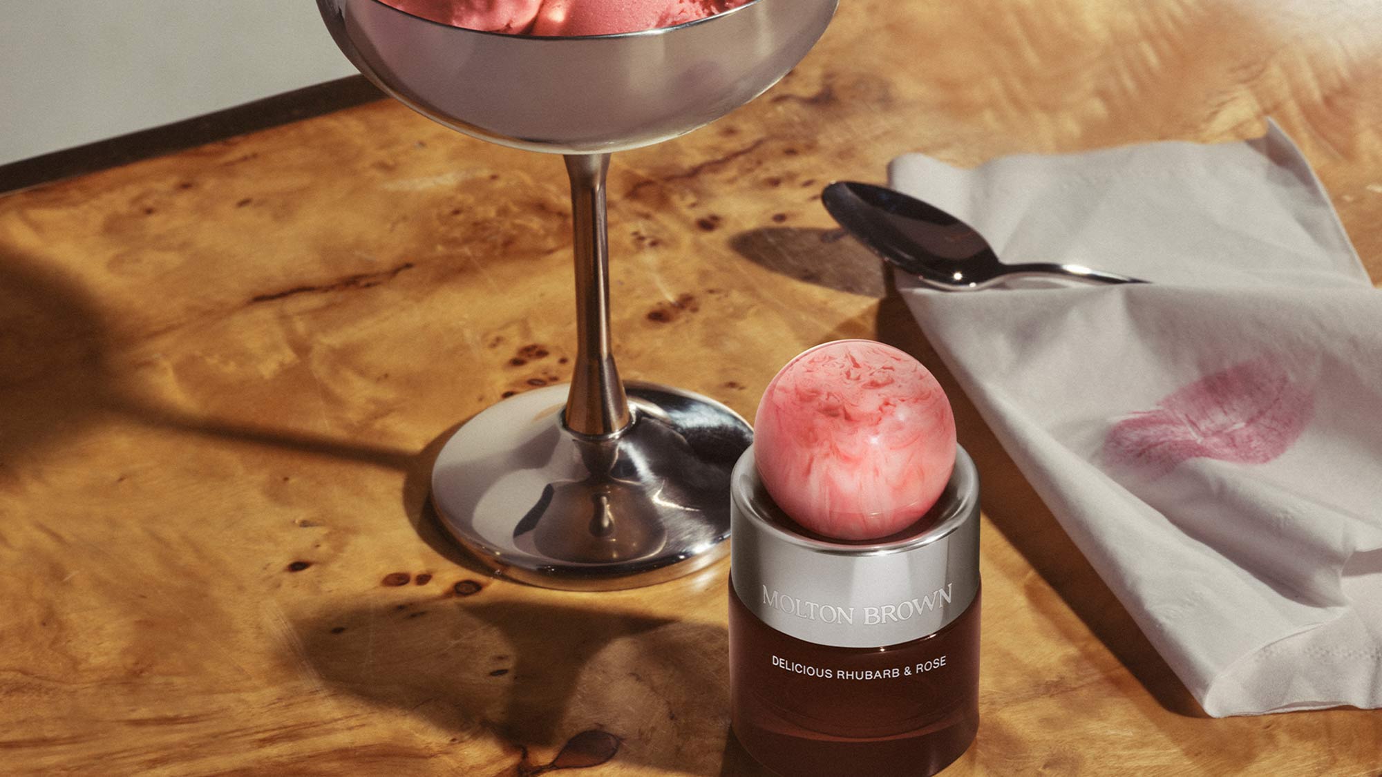 Molton Brown Delicious Rhubarb & Rosé fragrance bottle with pink ball applicator, displayed with cocktail and white cloth.