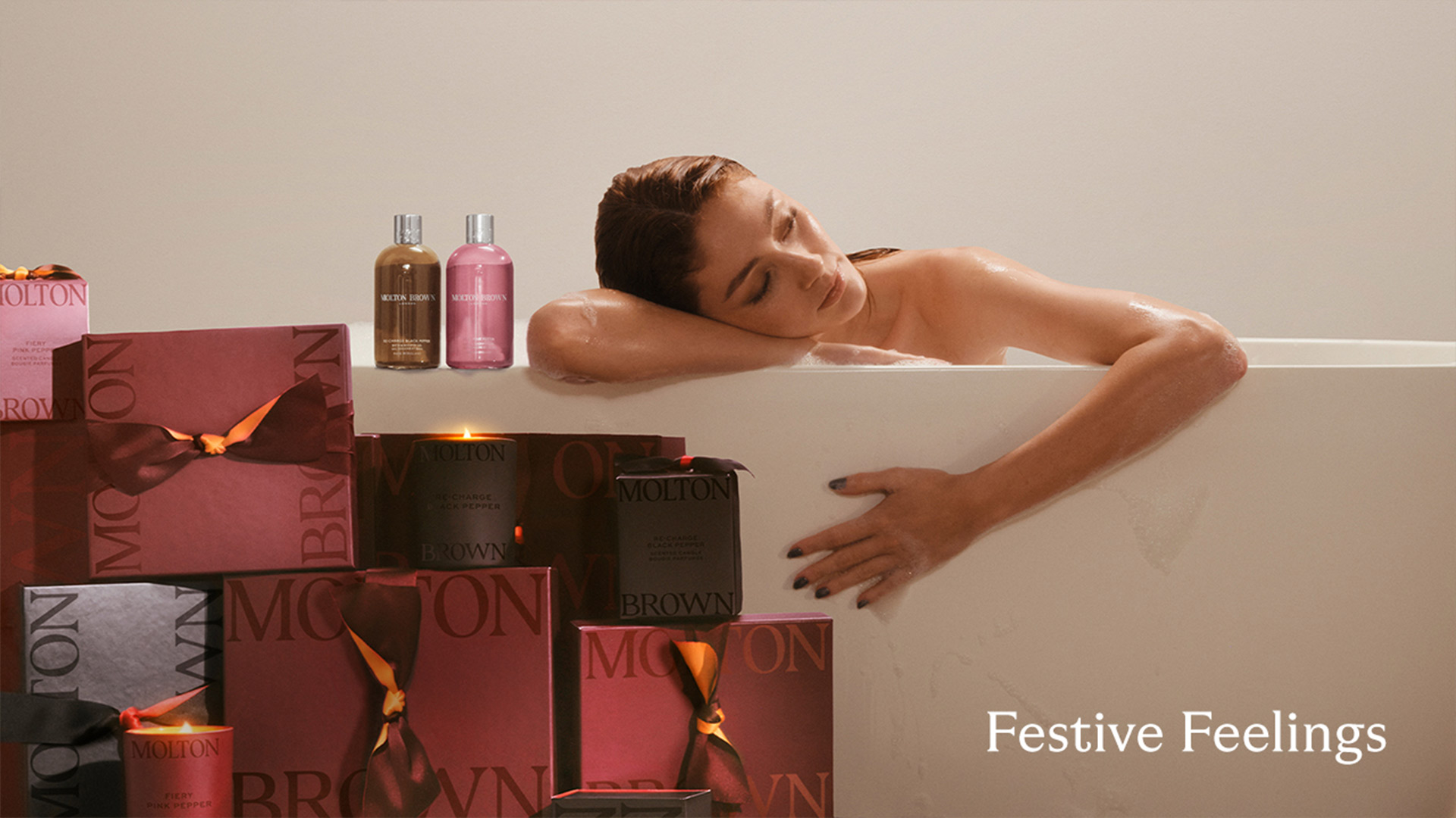 Woman relaxing in bath surrounded by Molton Brown gift boxes and toiletries in burgundy and gold packaging