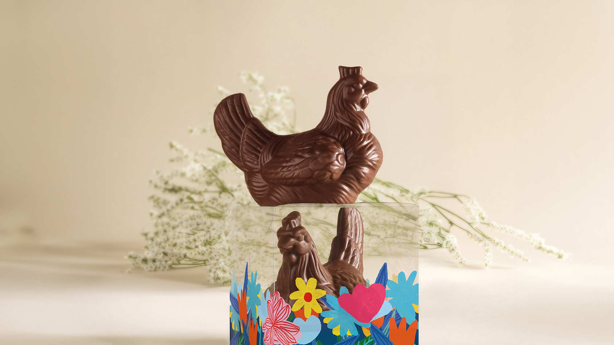 A chocolate hen on a box with colorful floral designs, set against a blurred light background with delicate white flowers.