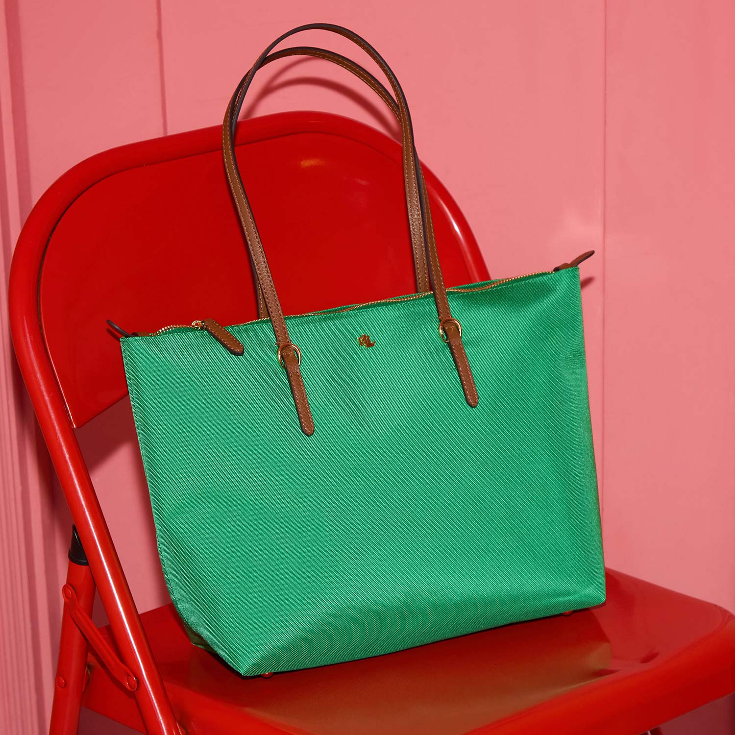 A green handbag with brown handles is placed on a red folding chair against a pink wall.