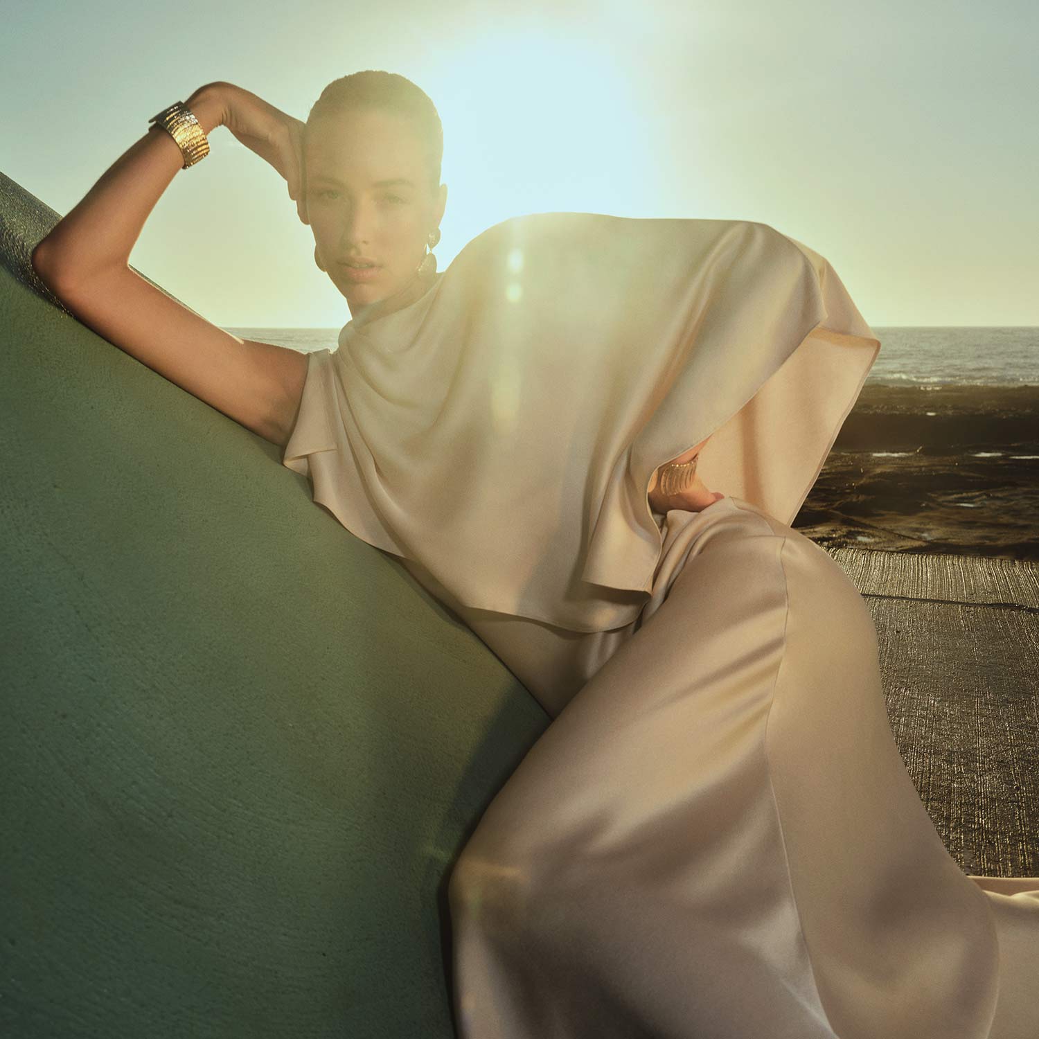 A person in an elegant, flowing outfit poses against a green structure with the sun setting in the background.