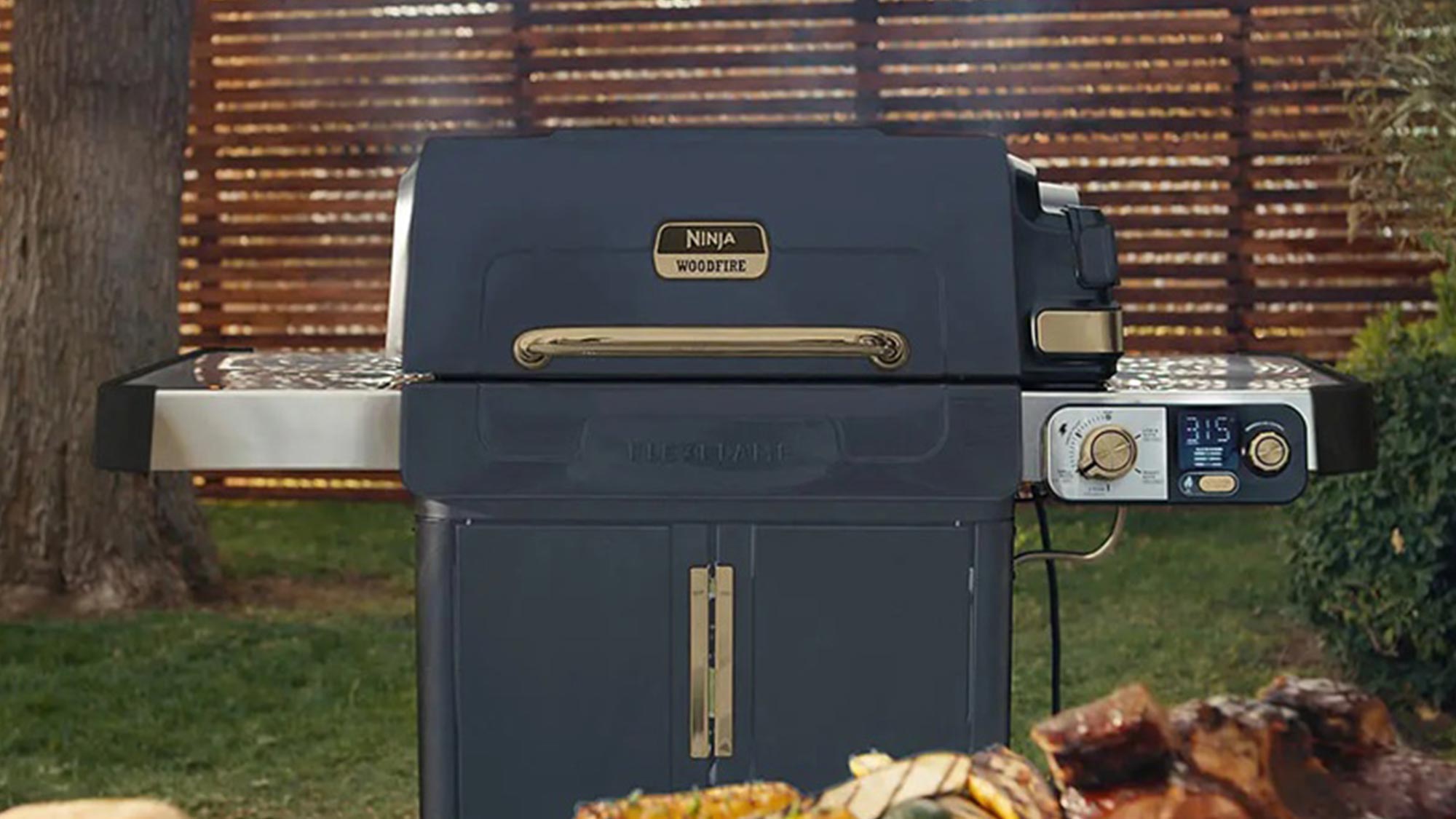 Navy Ninja Woodfire outdoor grill with control panel and grilled food, set in a garden with wooden fence backdrop