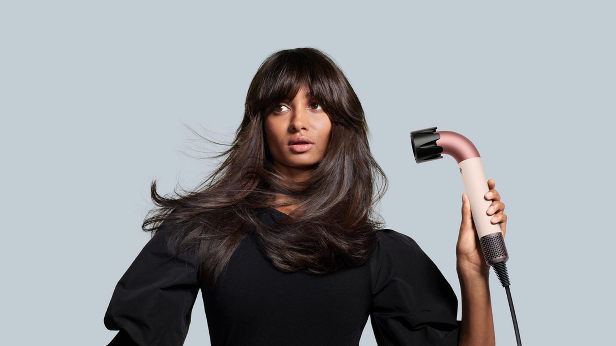 Woman with long dark hair holding a white and black hair styling tool against a grey background