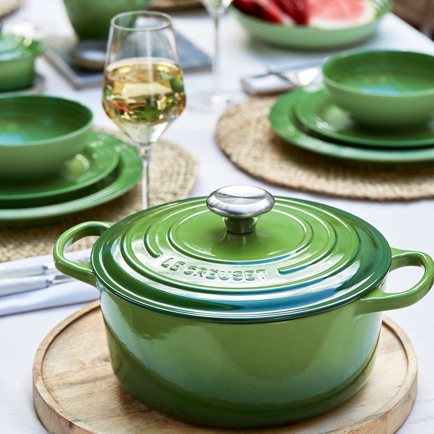 Green Le Creuset casserole dish with lid displayed on a table setting with matching green plates, natural placemats, and glassware