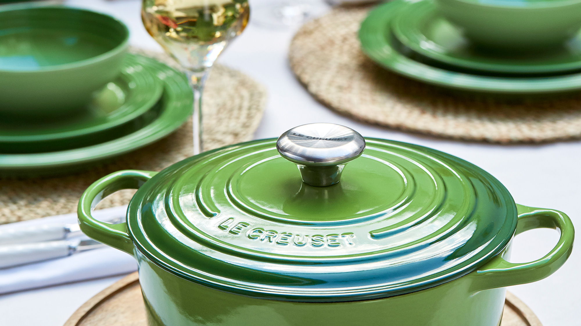 Green Le Creuset casserole dish with lid displayed on a table setting with matching green plates, natural placemats, and glassware