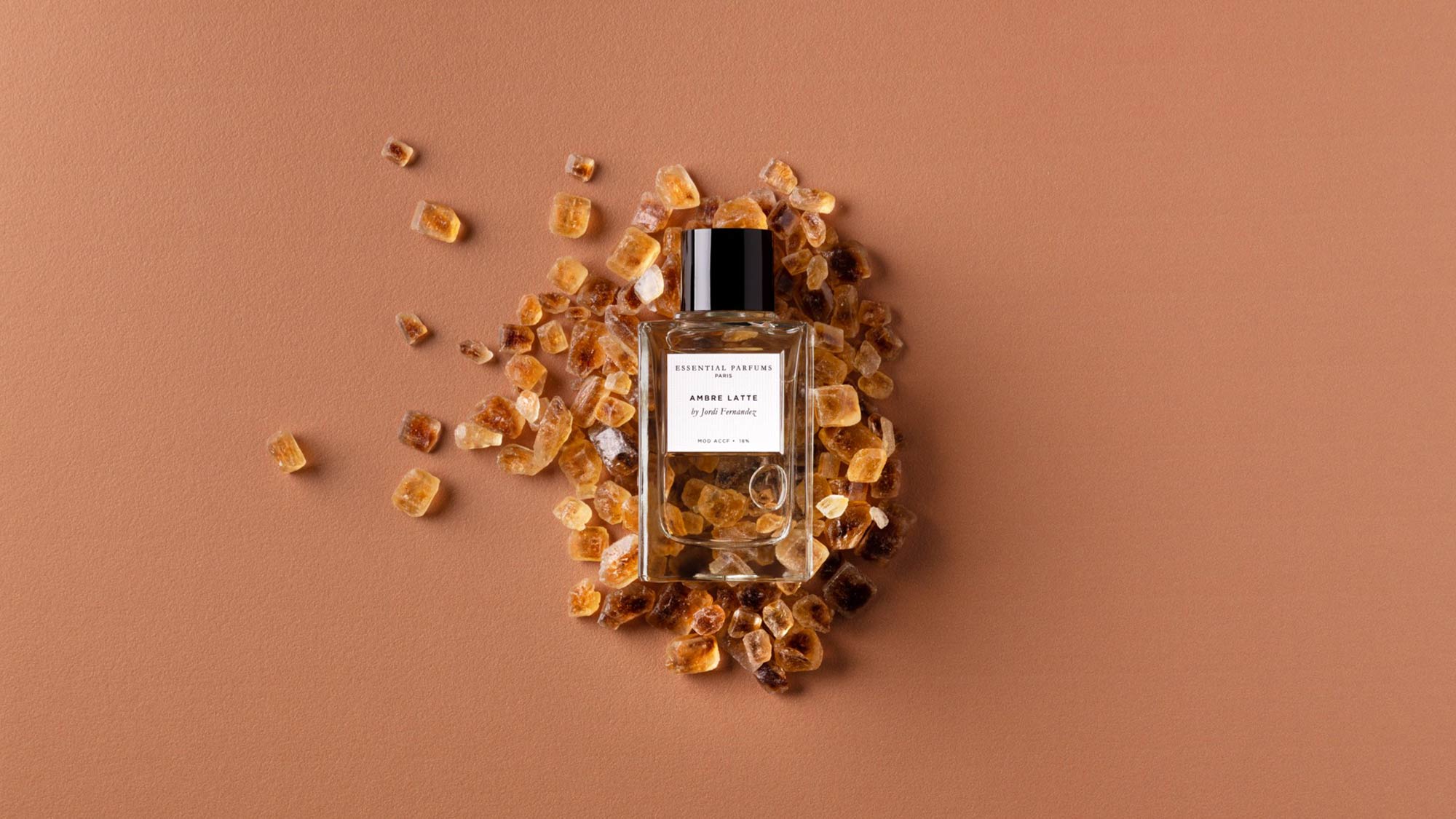 Essential Parfums Ambre Latte fragrance bottle surrounded by amber crystal pieces on neutral background