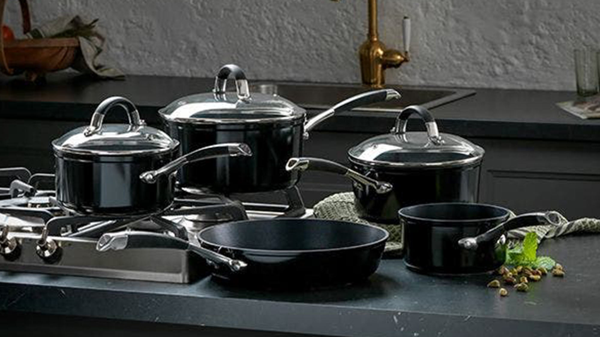 Black cookware set displayed on dark countertop, including pots with glass lids, frying pan, and cooking utensils