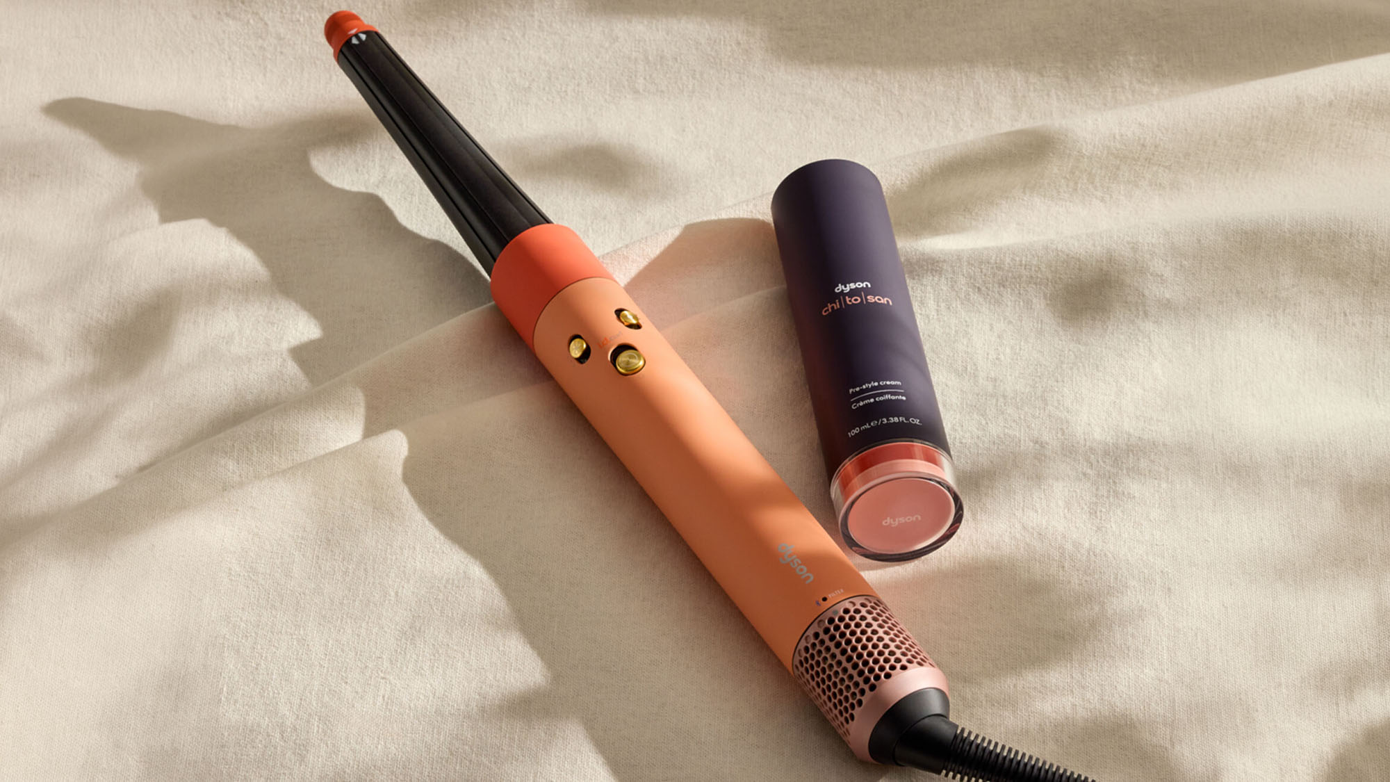 Coral-coloured hair styling tool with black handle and two beauty products arranged on cream textured fabric.