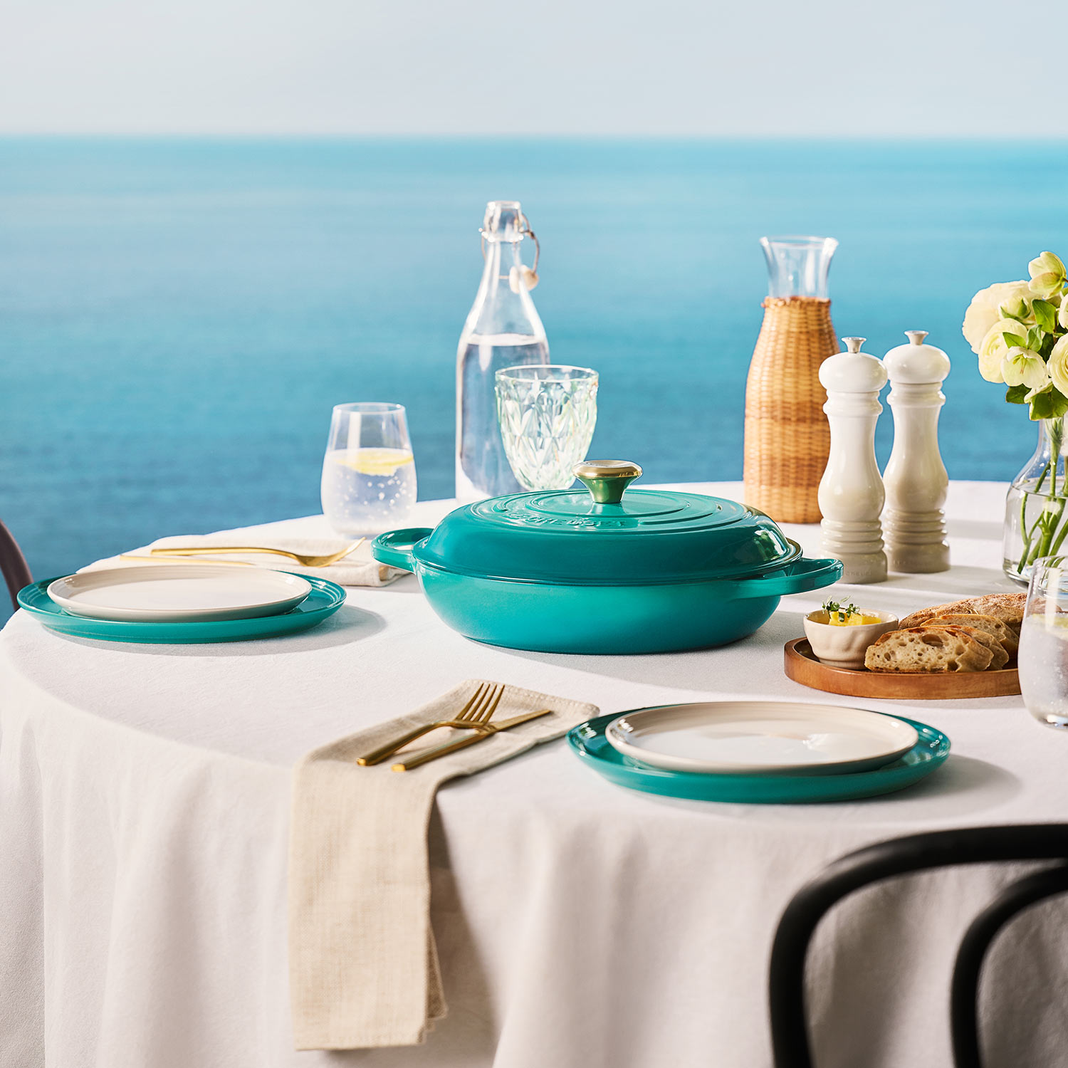 Turquoise cookware and tableware set on white table with water glasses, bread, flowers and condiments against blue background.