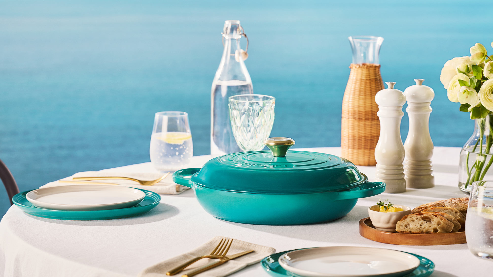 Turquoise cookware and tableware set on white table with water glasses, bread, flowers and condiments against blue background.