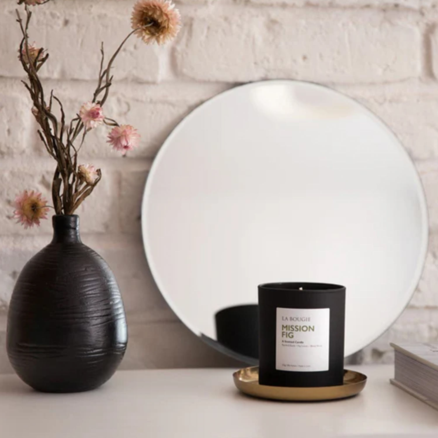 Black Mission Fig scented candle on brass tray beside dried flowers in dark vase and white circular mirror