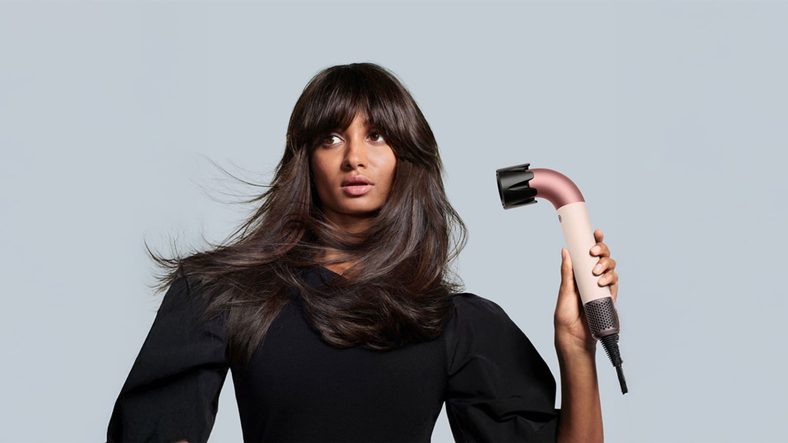 A woman with long hair holds a hairdryer against a plain background.