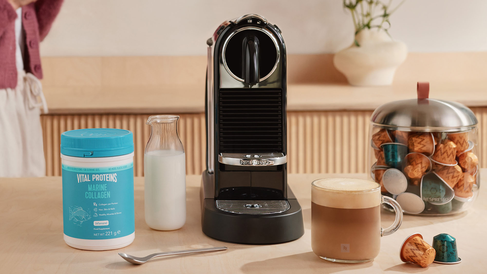 Coffee promotion showcasing a coffee machine, drinks, and collagen tub on a table, with a person in the background holding a mug.