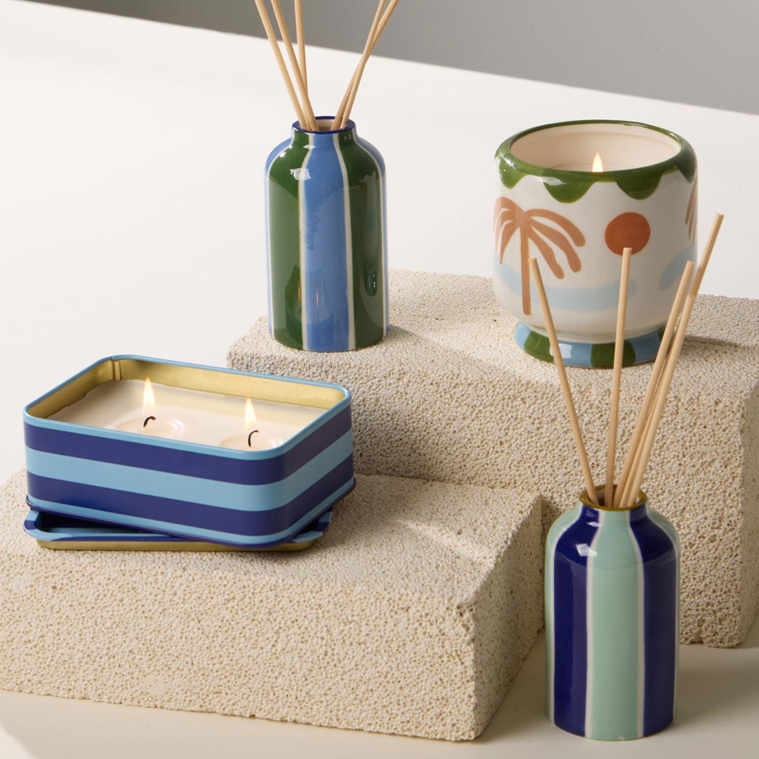 Striped reed diffusers and colourful candles are placed on textured stone blocks. The items feature a mix of blues, greens, and bold patterns, with some candles lit, emitting a soft glow.