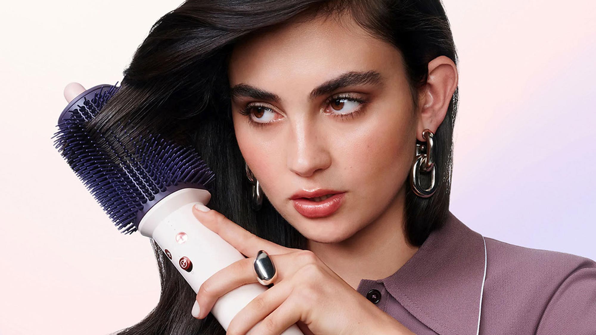 Woman using Dyson hair styling brush with blue bristles on dark hair against light gradient background