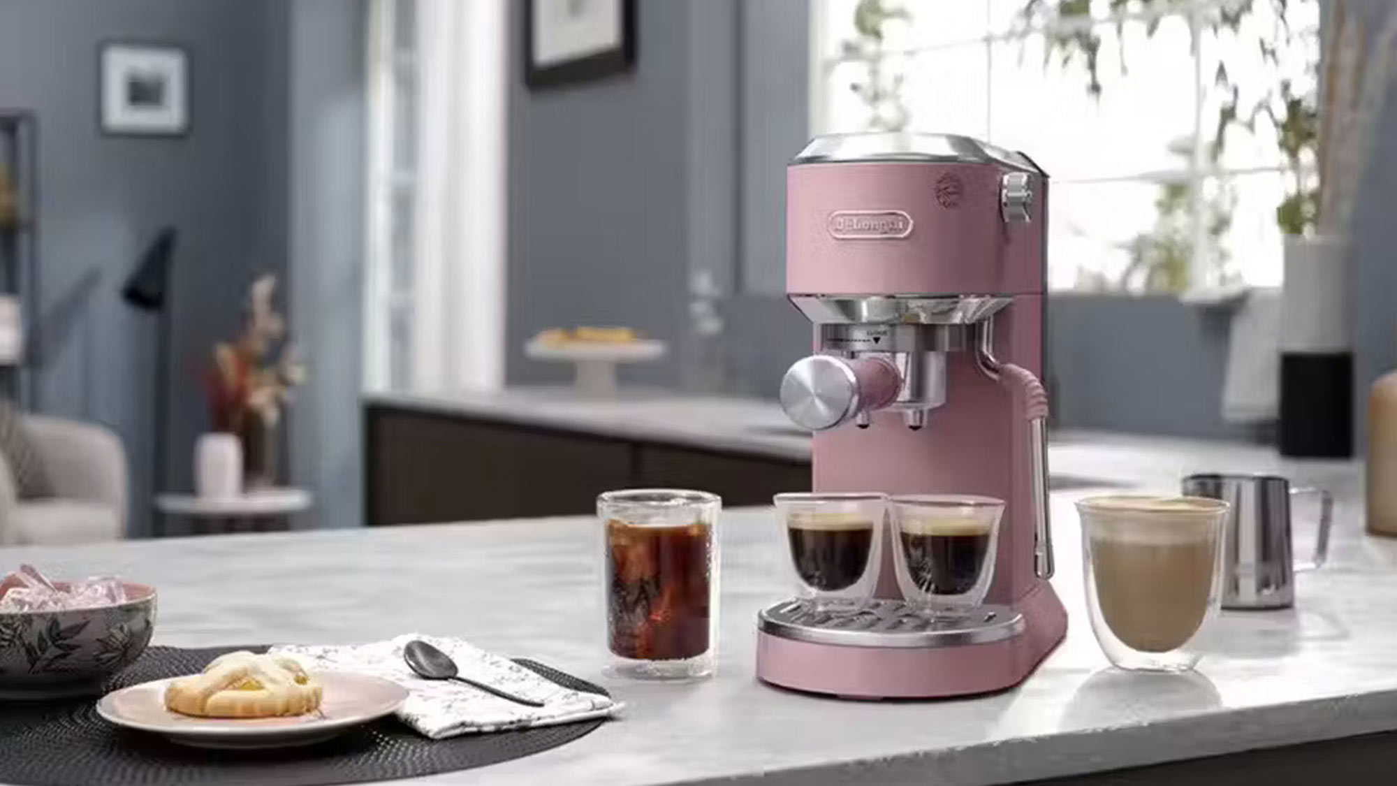 A pink espresso machine on a countertop with various drinks and pastries nearby in a modern kitchen setting.