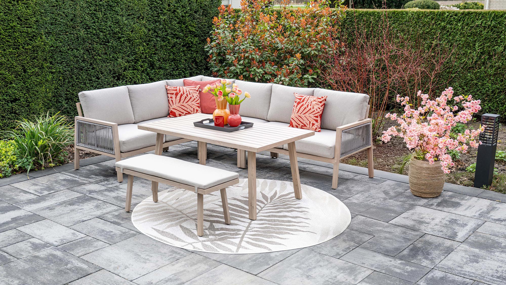 A patio set with a corner sofa, table, and bench on gray tiles, decorated with pink flowers and cushions, surrounded by greenery.