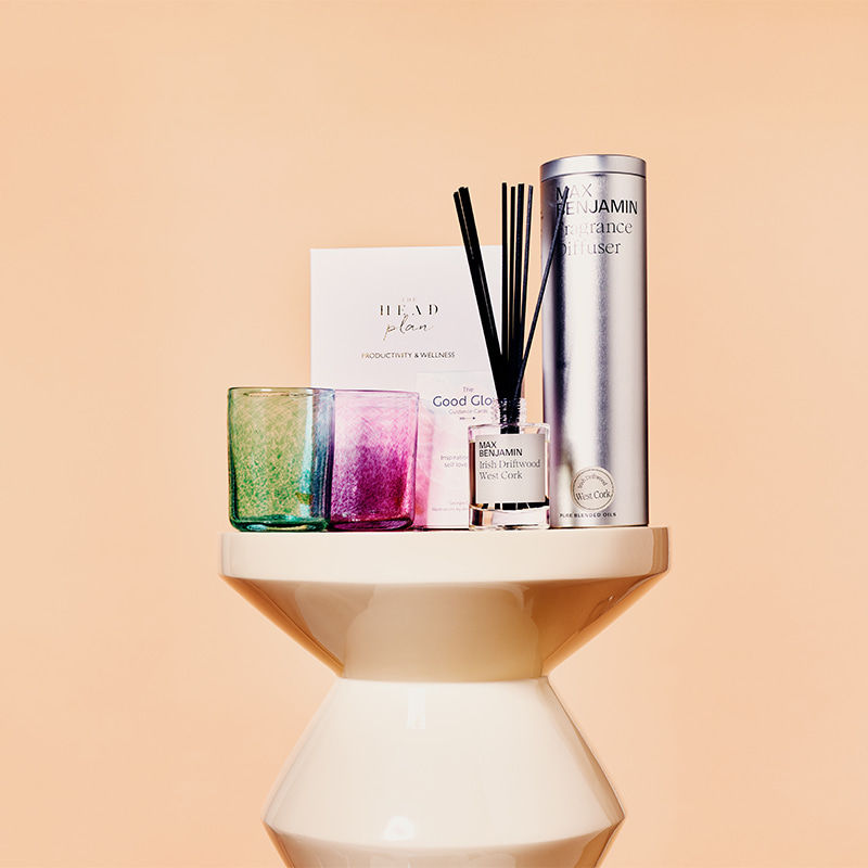 A set of colorful candles, a fragrance diffuser, and a box are arranged on a round pedestal against a peach background.
