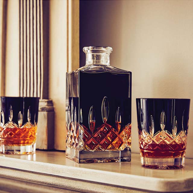 A decanter filled with dark liquid and two matching glasses with amber liquid stand on a shelf in an elegant setting.