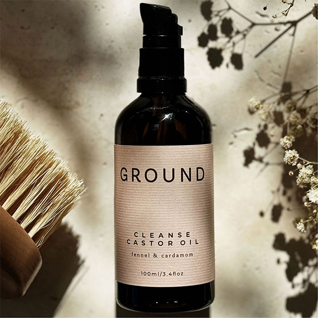 A bottle of GROUND cleanse castor oil with fennel and cardamom, placed next to a brush and white flowers.