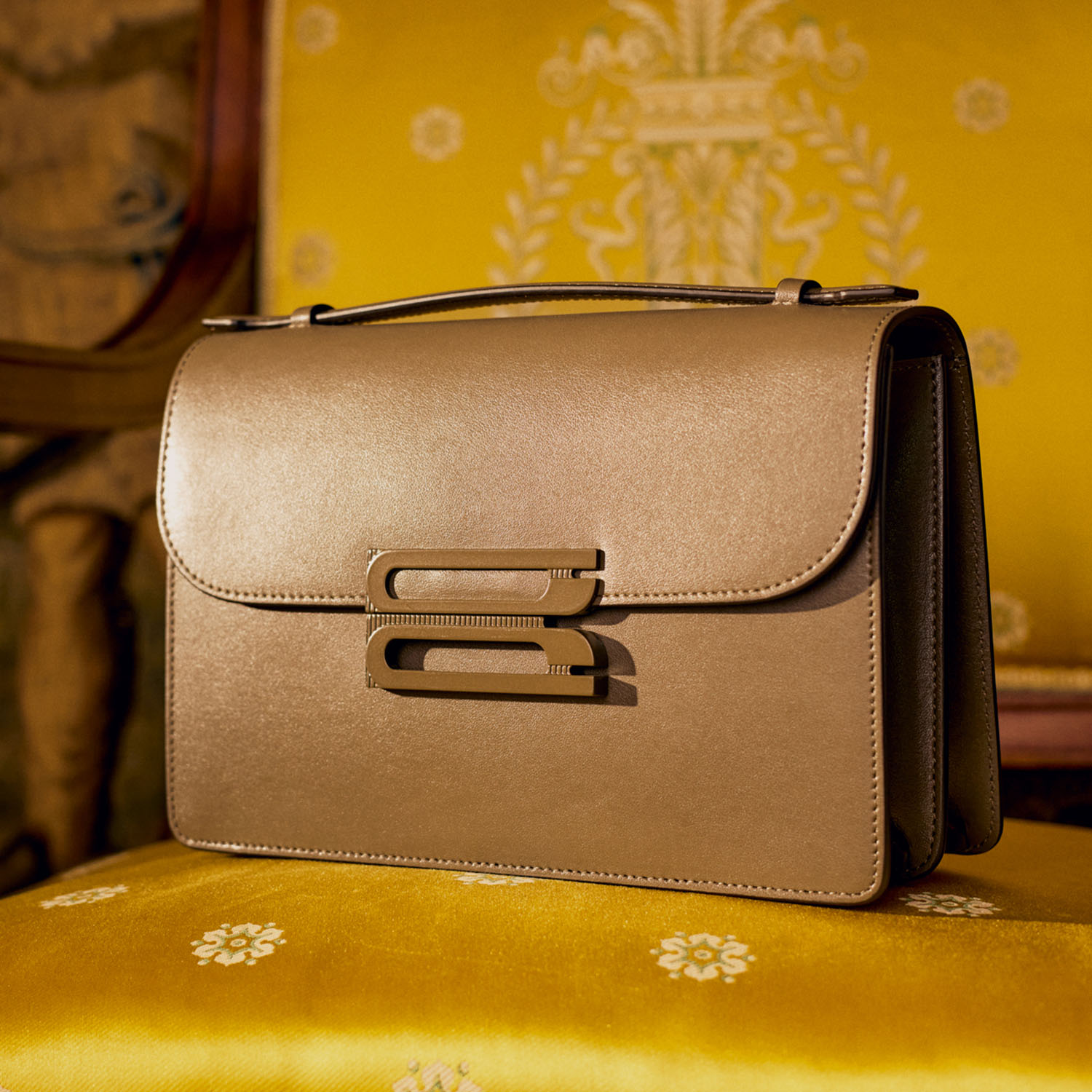 A beige handbag with a modern clasp design sits on a patterned yellow surface with ornate furniture in the background.