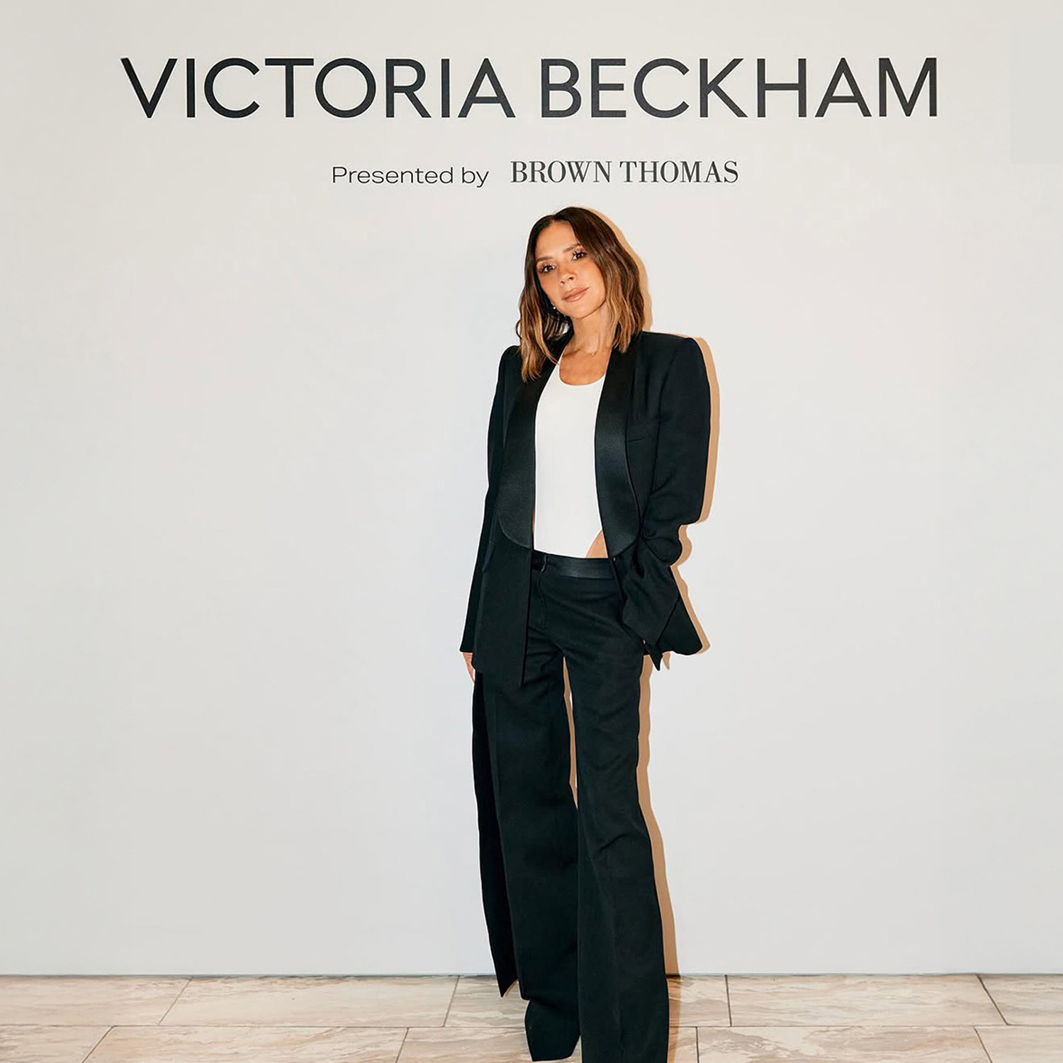 A woman in a black suit and white top stands in front of a 'Victoria Beckham' sign.