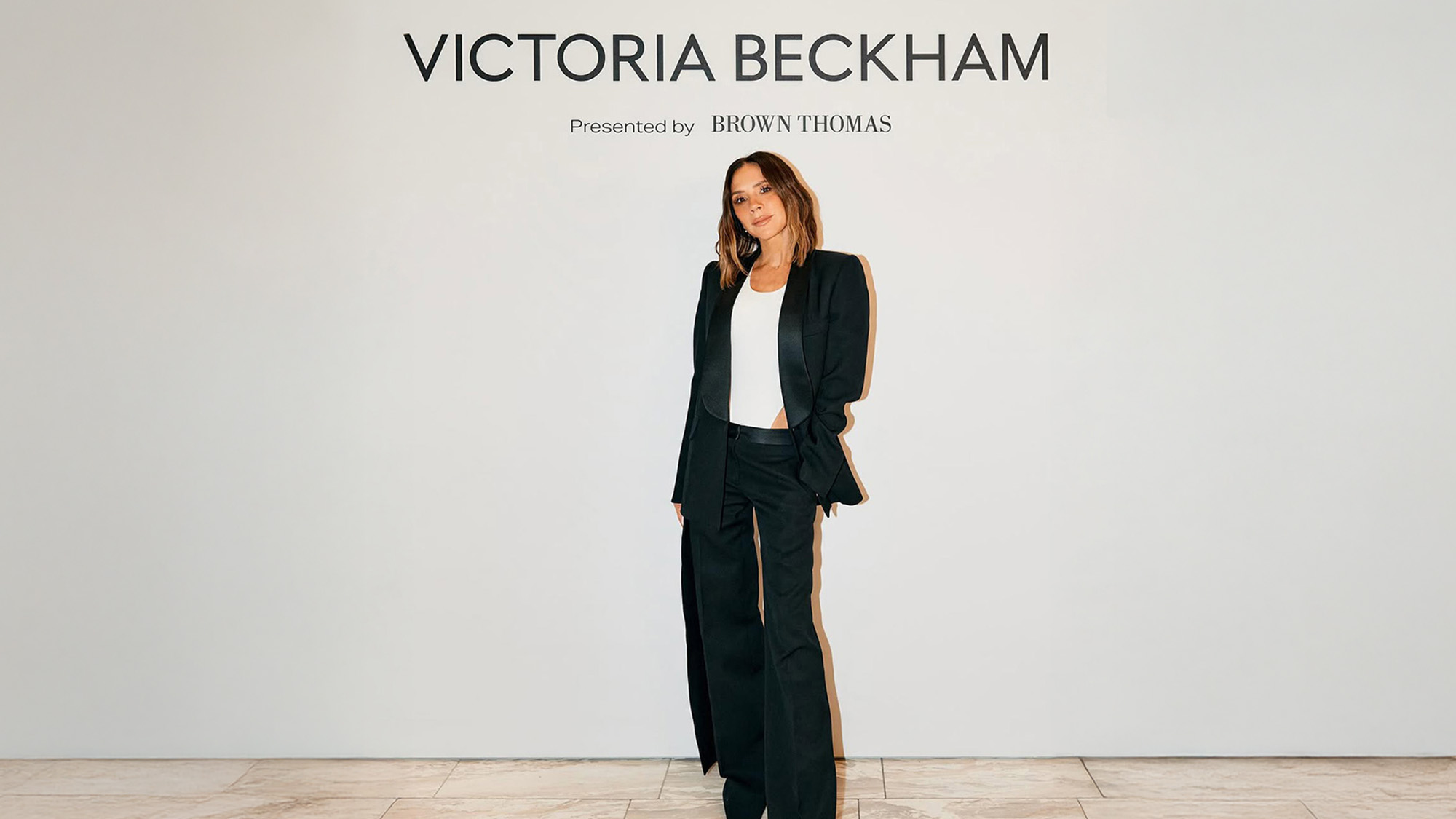 A woman in a black suit stands under a 'Victoria Beckham' sign presented by Brown Thomas.