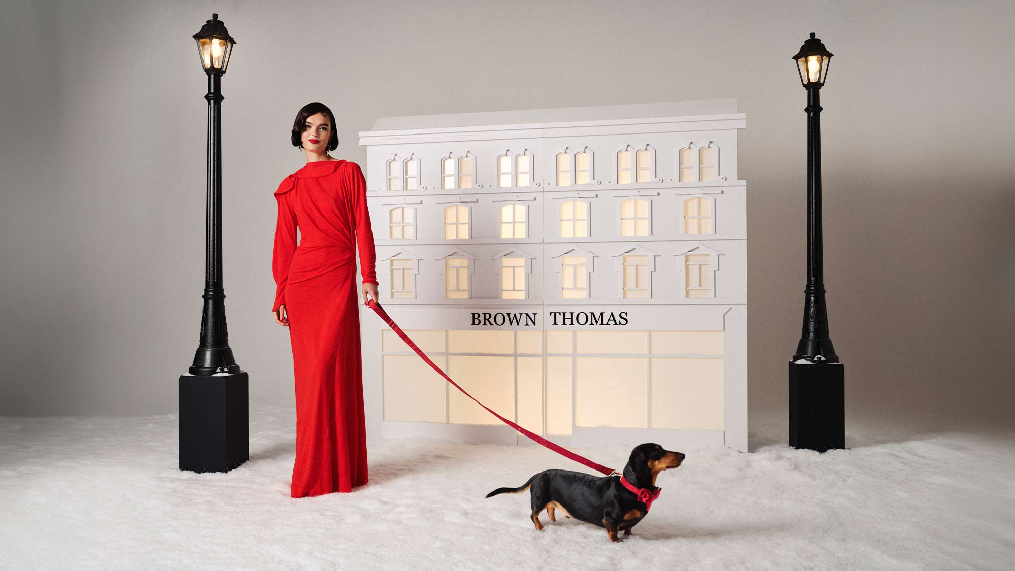 A man in a tuxedo and a woman in a dark dress stand in a snowy scene with street lamps and a 'Brown Thomas' backdrop.