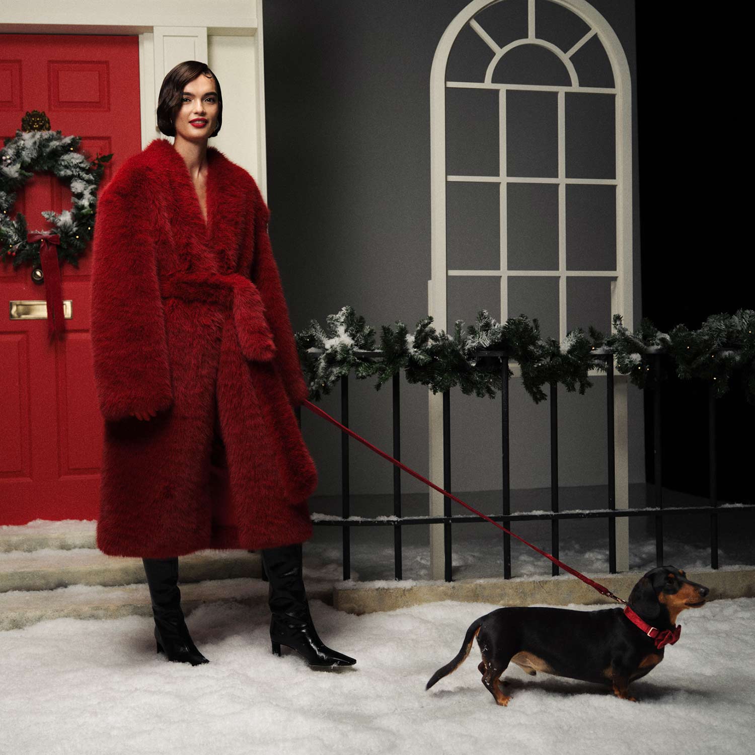 A woman in a red coat stands in snow outside a house with a red door, walking a small dog on a red leash.