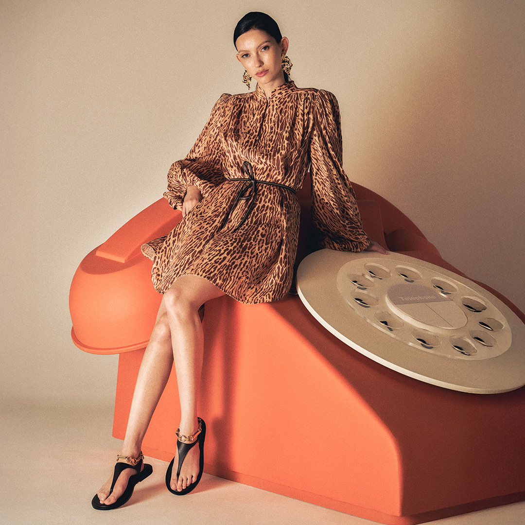 A woman in a leopard-print dress sits on a large, orange, telephone-shaped chair.